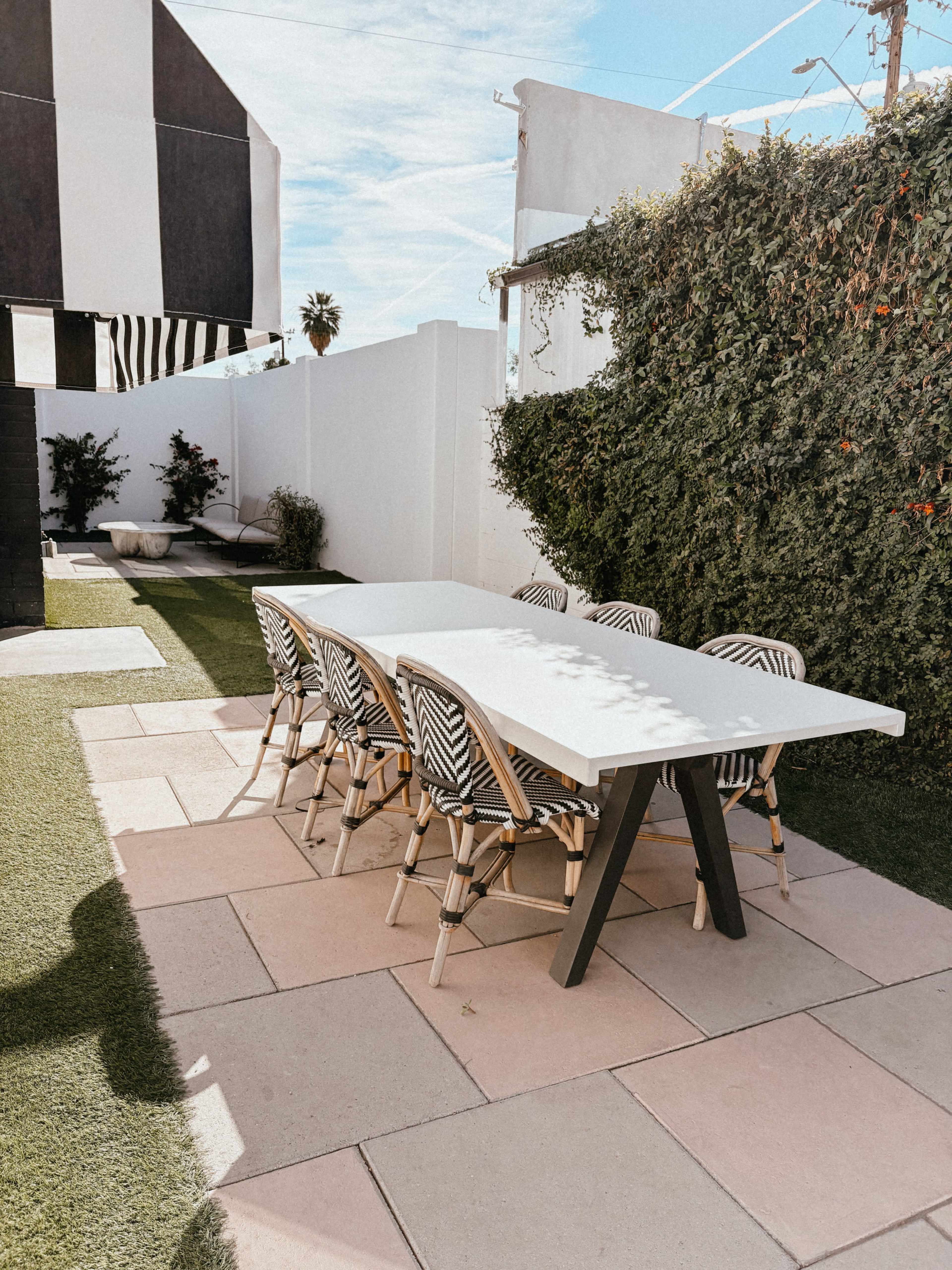 A modern outdoor dining table with striped chairs is set on a patio surrounded by greenery and soft landscaping.