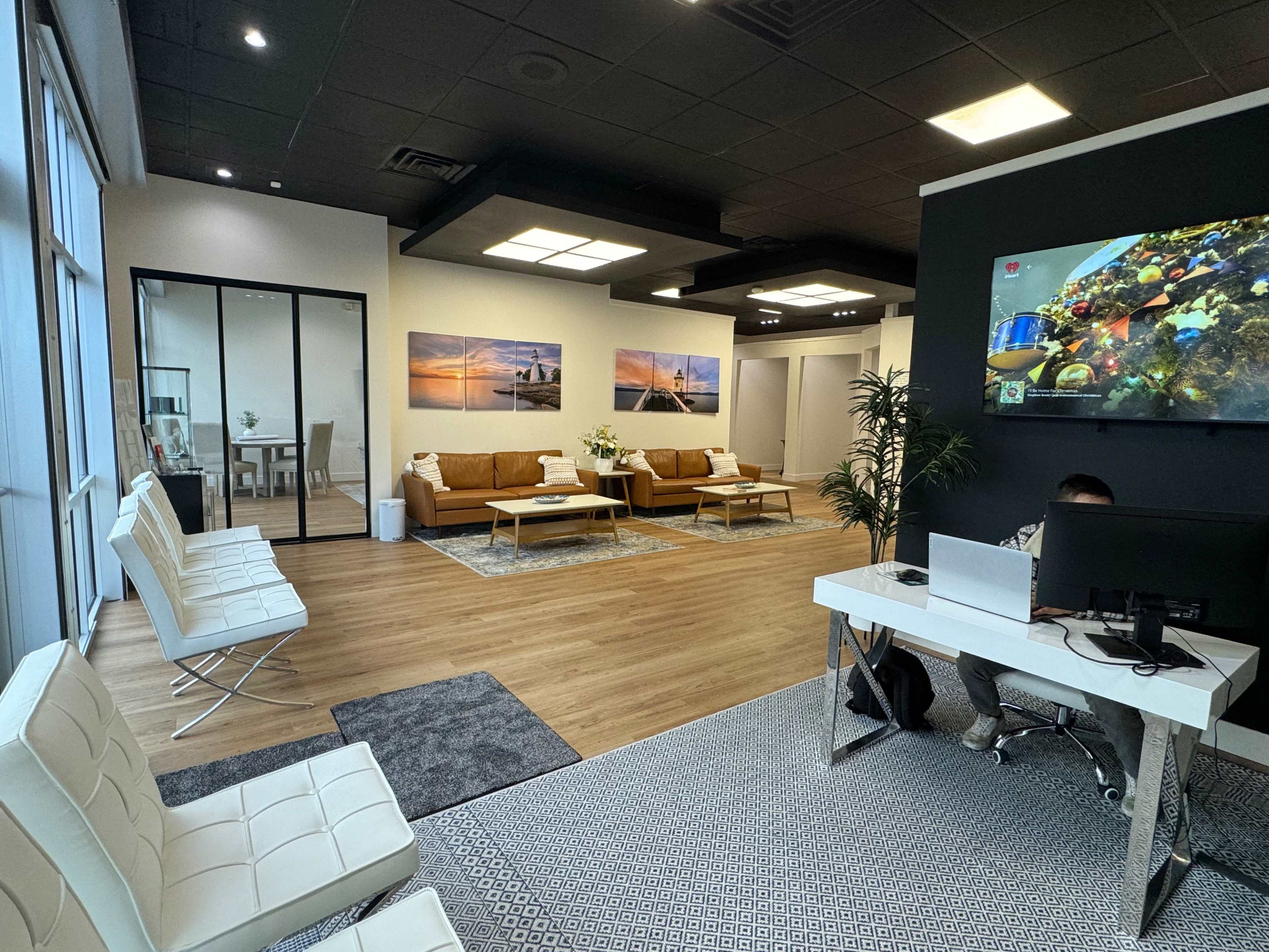 A modern office reception area featuring two brown sofas, white chairs, a large flat-screen TV on the wall, and wood flooring.