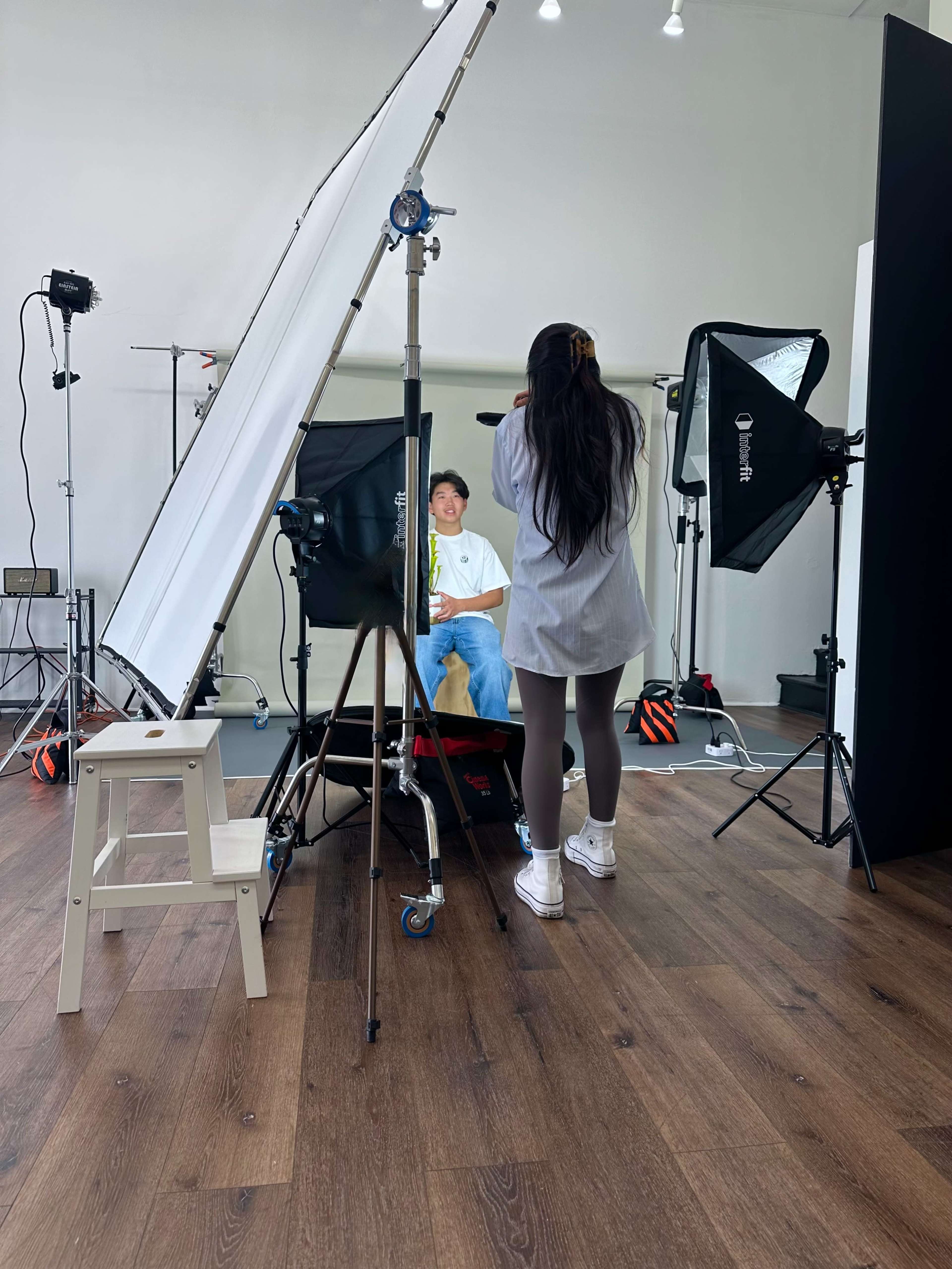 A photographer is capturing a portrait of a person sitting in a studio environment with various lighting and backdrop equipment.