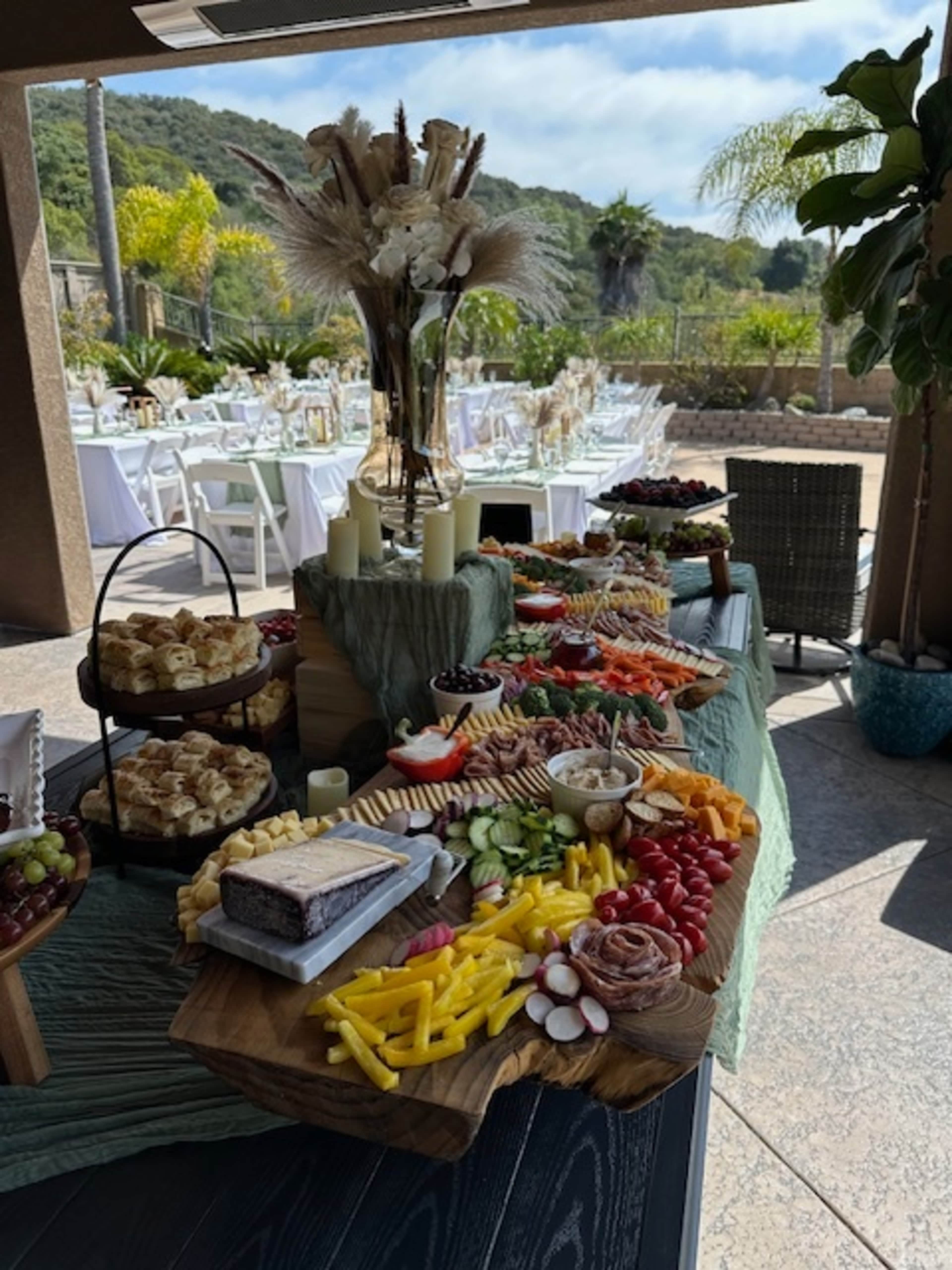 A large wooden platter displays an assortment of cheeses, meats, fruits, and vegetables, set up on a table at an outdoor venue with greenery in the background.