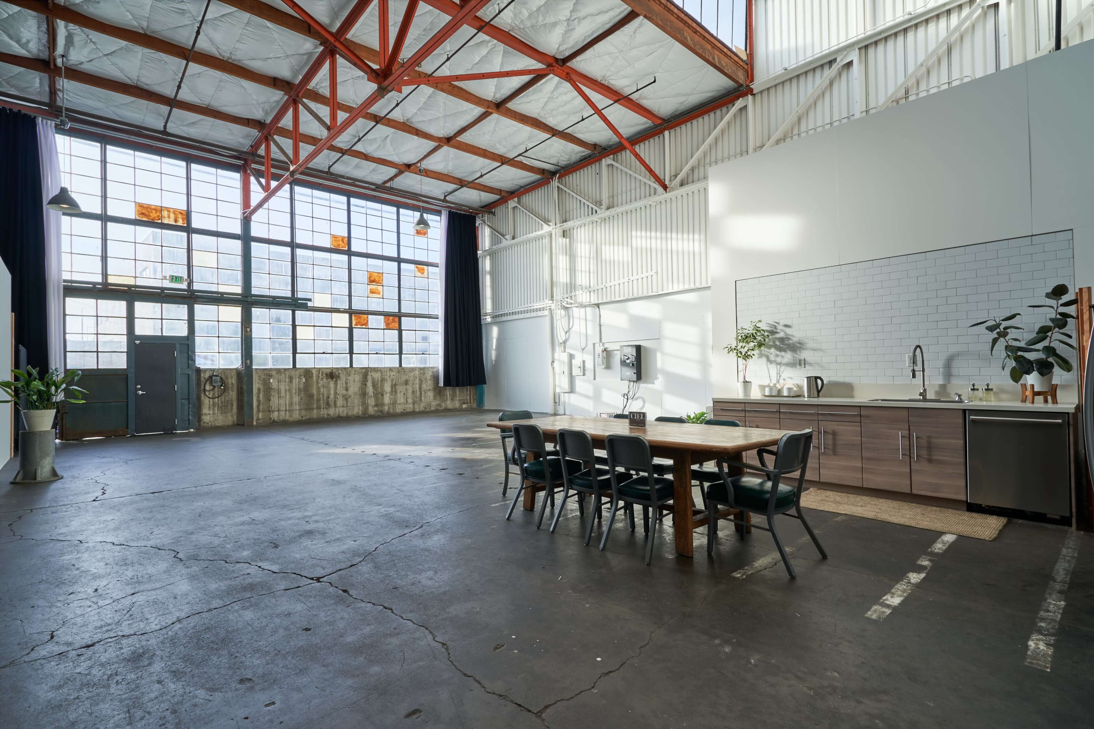 A spacious industrial-style room features large windows, a wooden table with chairs, and a kitchen area along one wall.