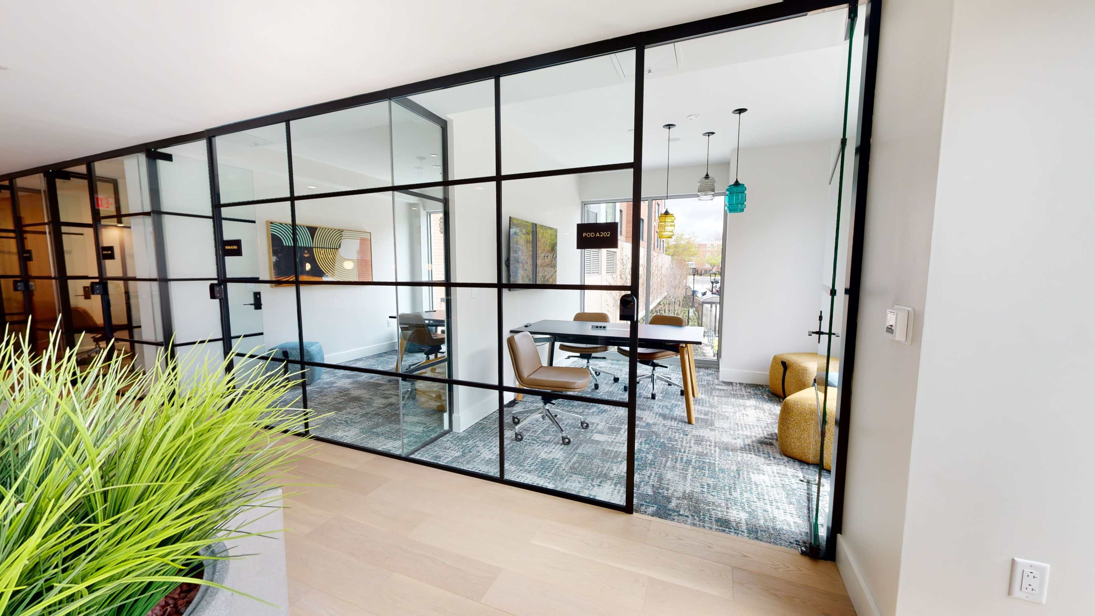 A modern office space is divided by glass walls, featuring a meeting area with a conference table and chairs, and accented by green plants outside.
