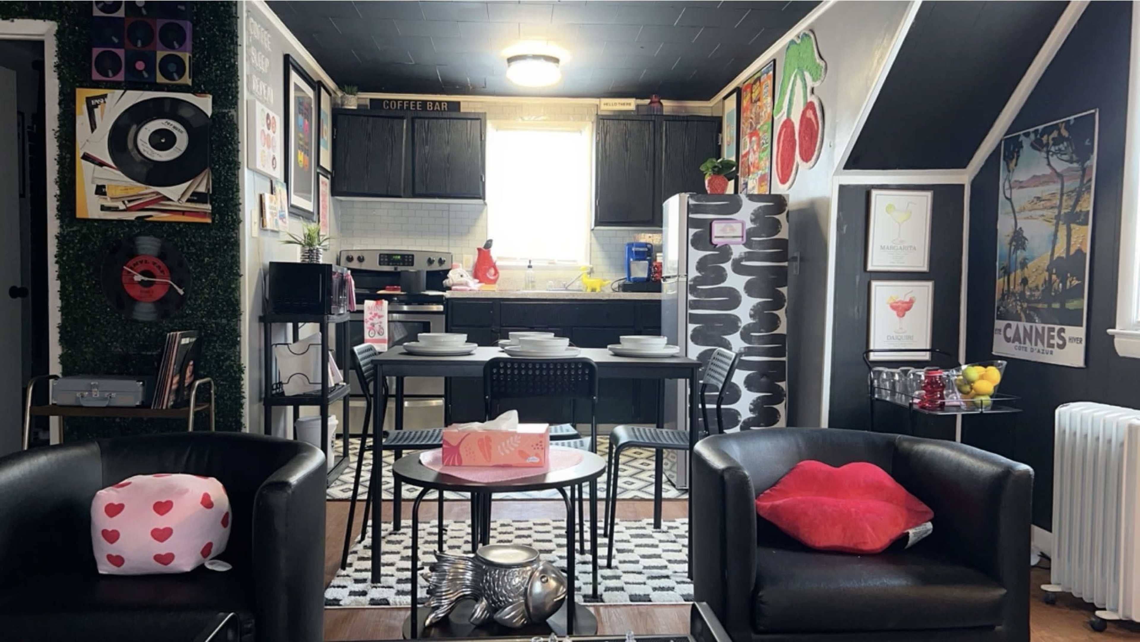 The image shows a stylish living space featuring a kitchen with a coffee bar, black cabinets, a dining area with a round table, and decorative art on the walls.