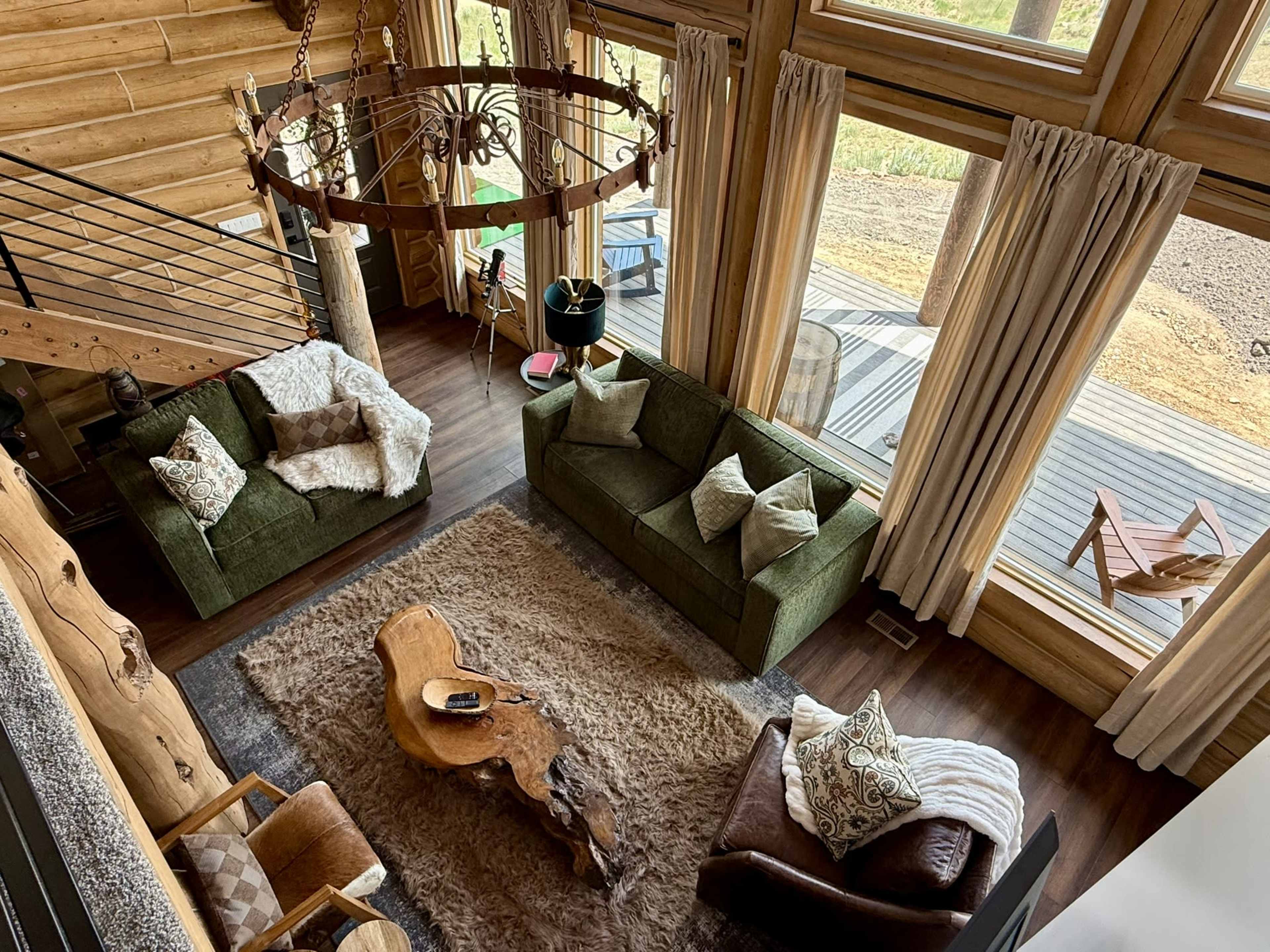 The image shows a spacious wooden interior living room featuring two sofas, a coffee table made from a tree trunk, and large windows providing natural light.
