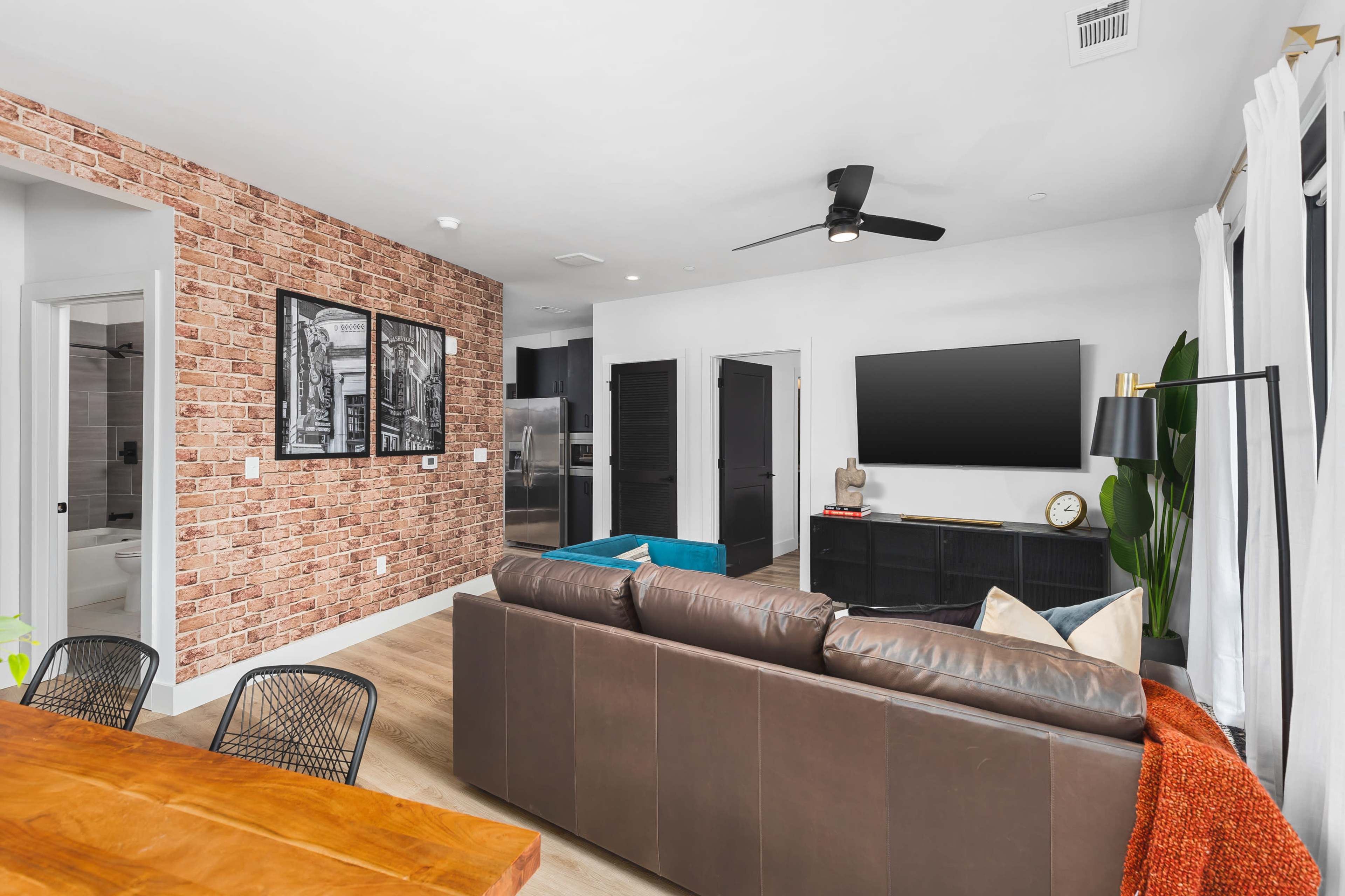 A modern living space featuring a brown leather sofa, a dining table, exposed brick wall art, and a wall-mounted television.