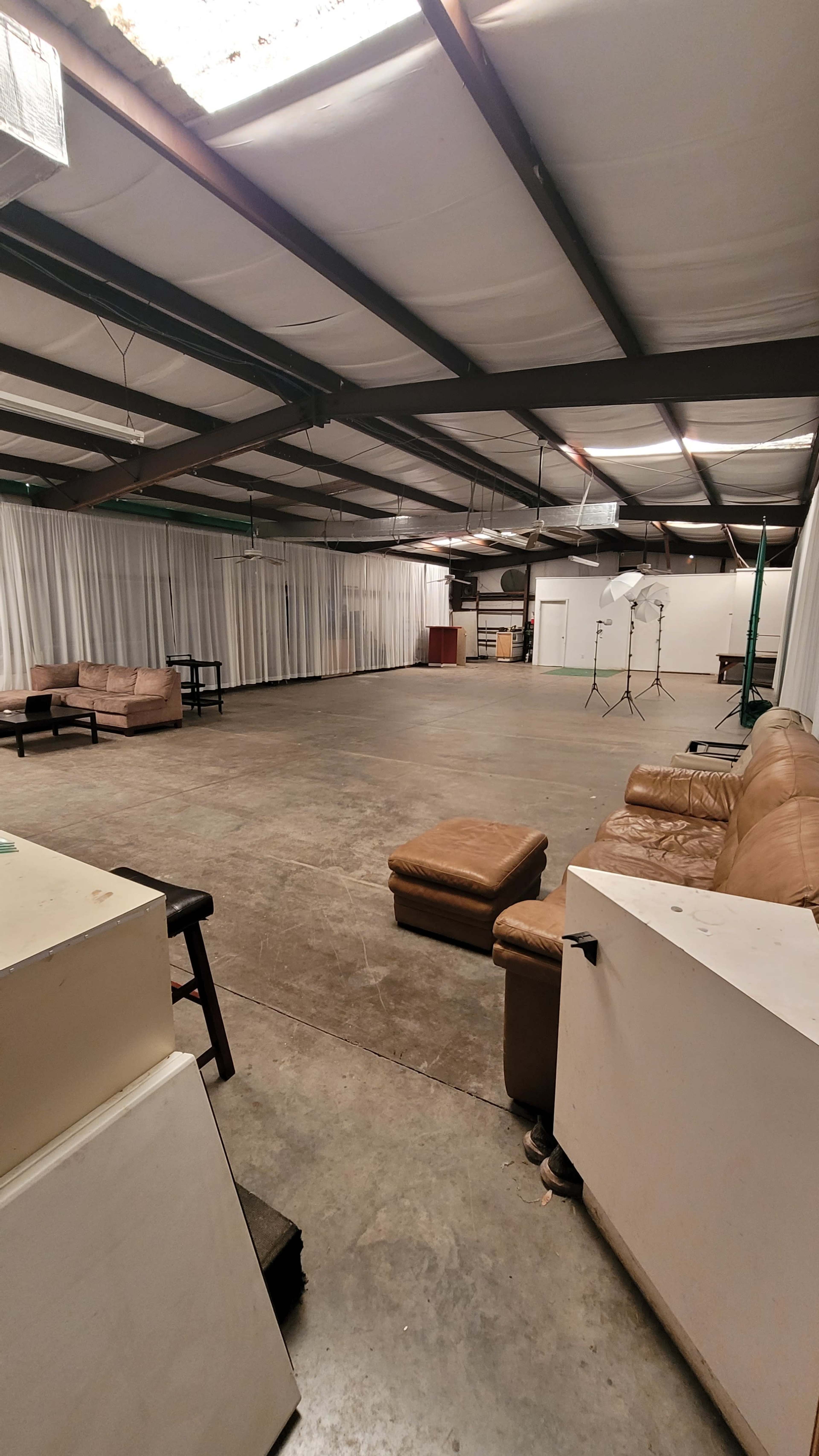 The image shows a spacious industrial room with a concrete floor, scattered furniture, and white curtains along the walls.