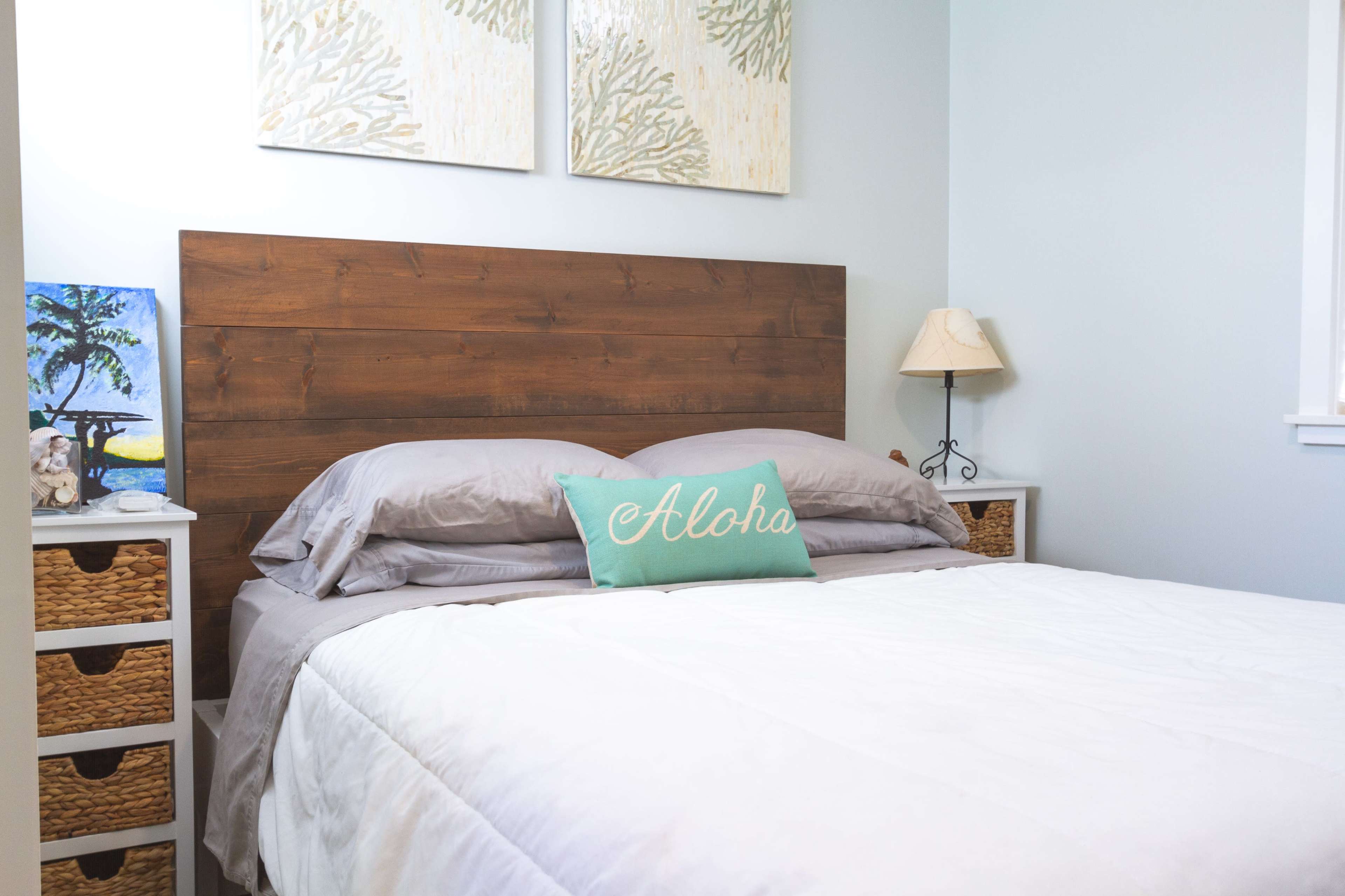 A neatly arranged bedroom with a wooden headboard, gray bedding, and a decorative pillow that reads "Aloha."