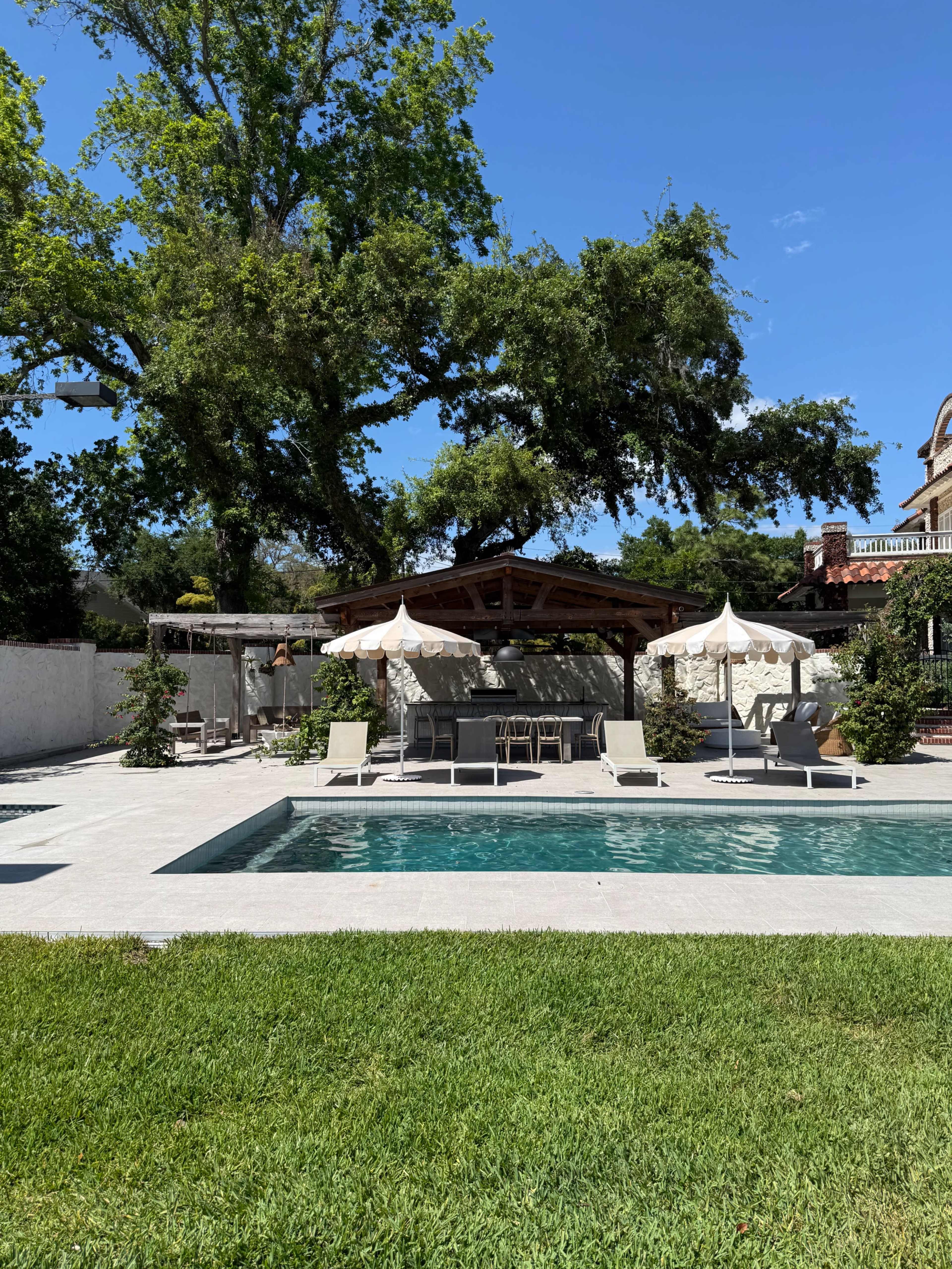 A serene outdoor scene features a swimming pool with lounging chairs and a shaded patio surrounded by greenery.