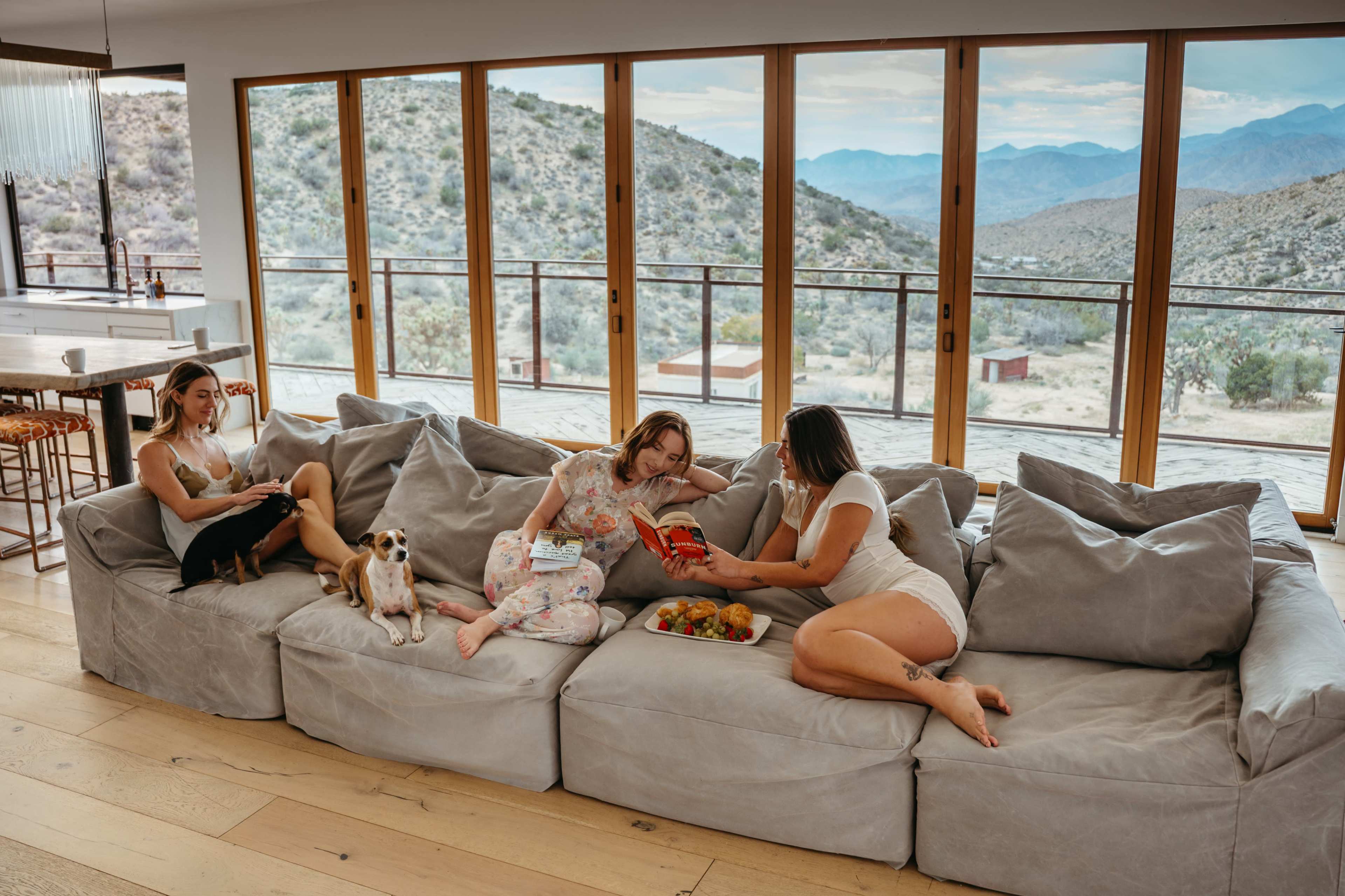 Three women are seated on a large sectional sofa in a modern, open living space that overlooks a mountainous landscape, while a small dog rests nearby.