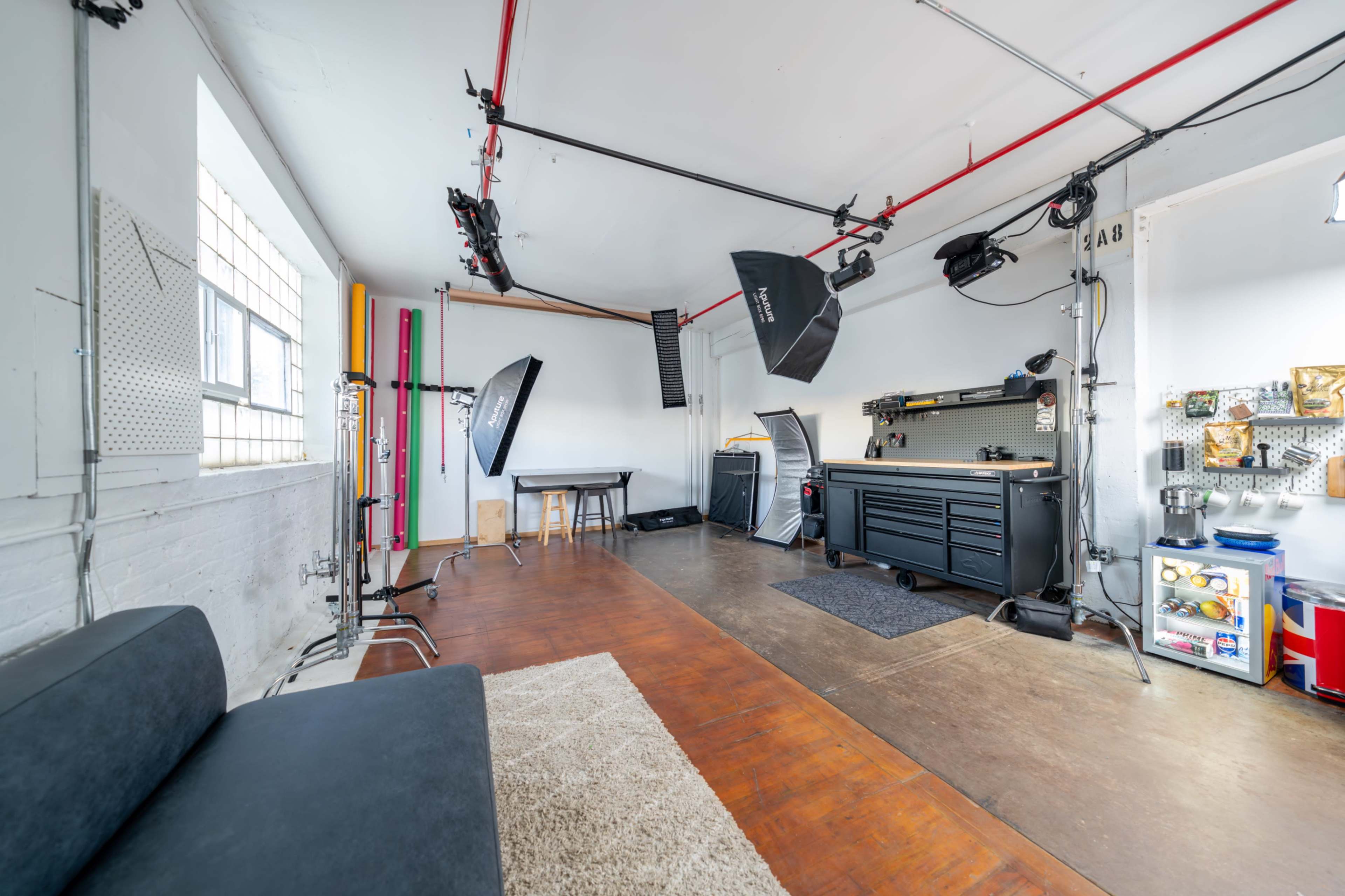 The image shows a spacious photography studio with various lighting equipment, a workbench, and a lounge area featuring a couch.
