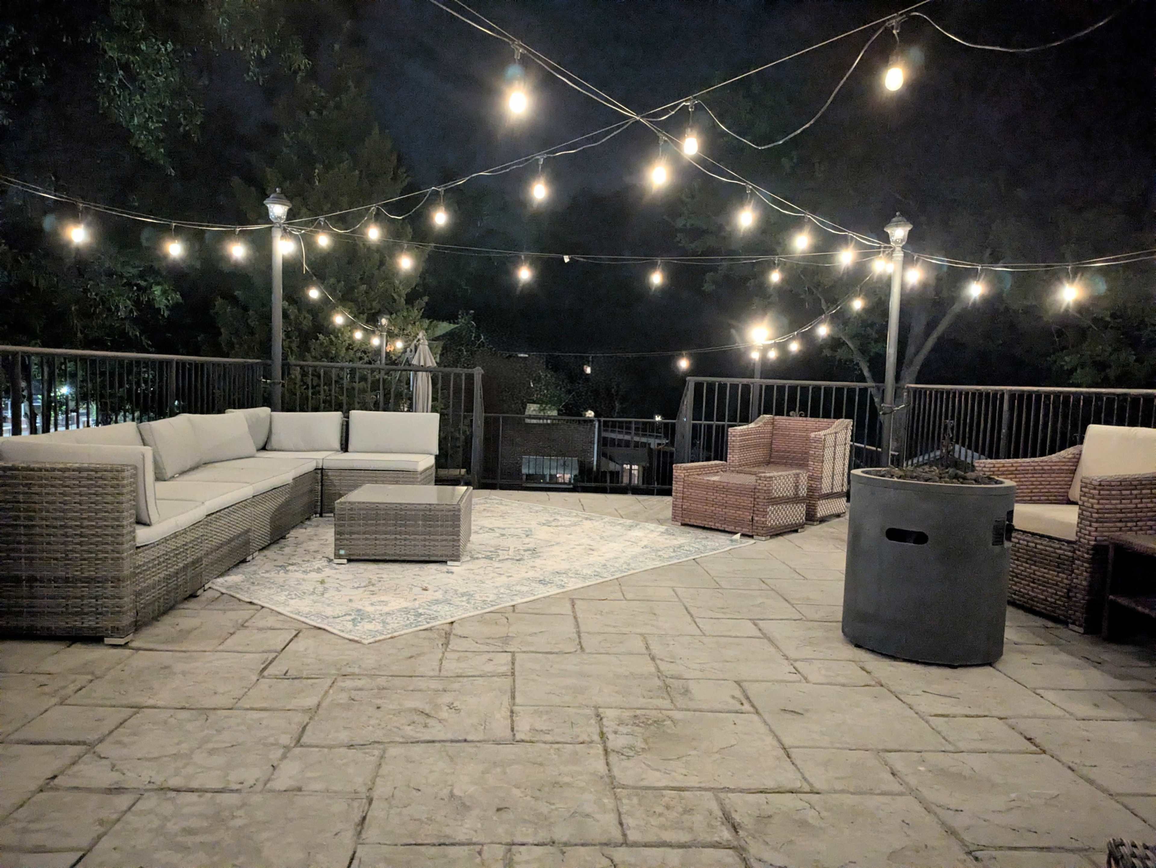 A landscaped outdoor seating area at night is illuminated by string lights, featuring wicker furniture and a fire pit.