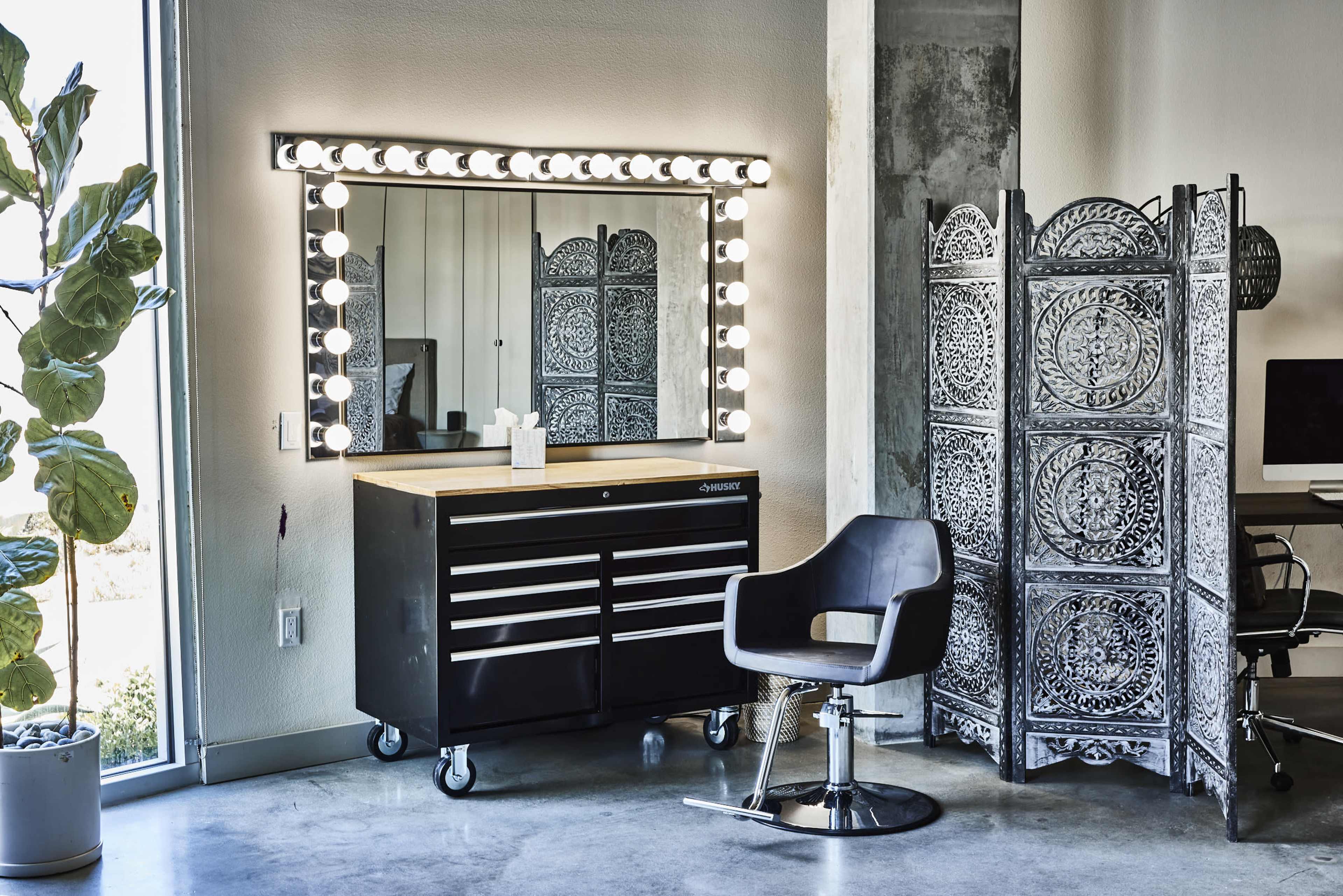 The image shows a modern salon setup featuring a large illuminated mirror, a cosmetic tool chest on wheels, a black styling chair, and a decorative privacy screen.