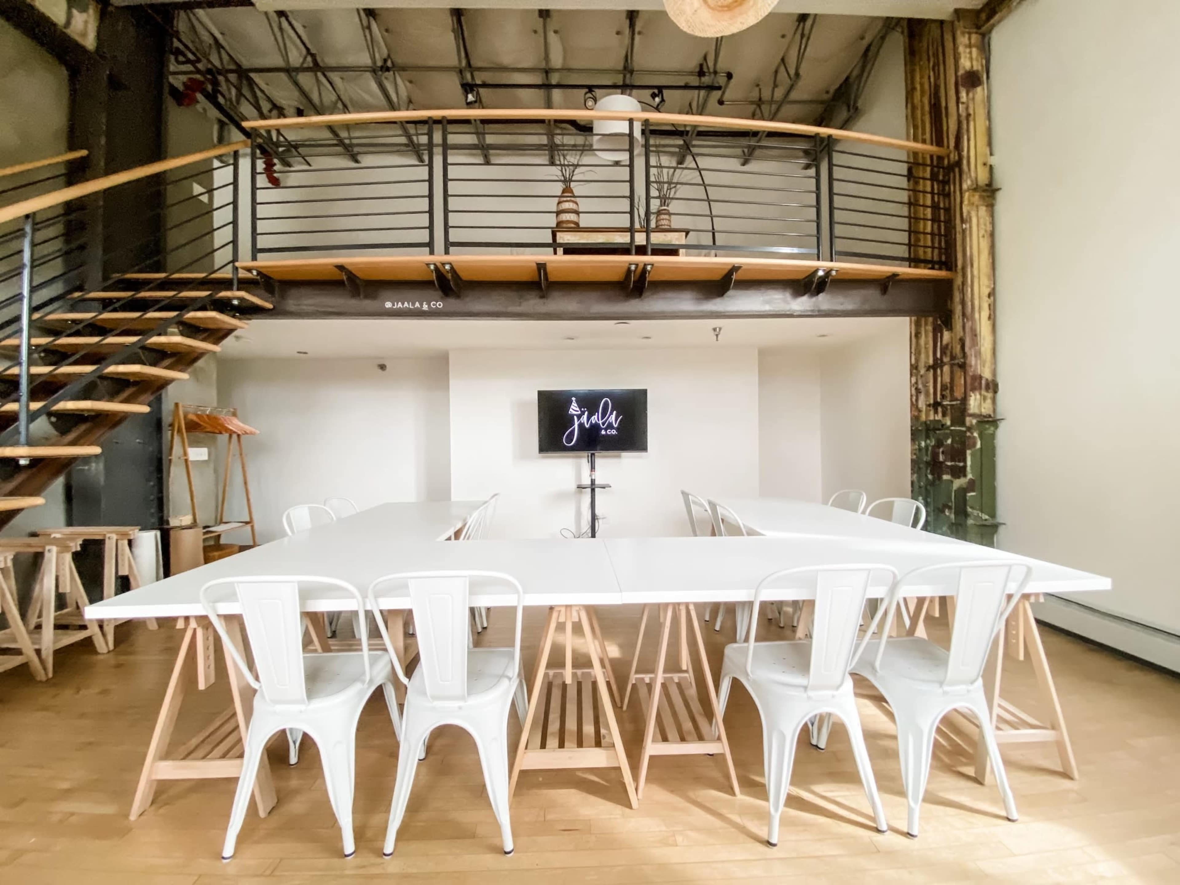 A spacious meeting room with a long white table surrounded by metal chairs, a display screen on a stand, and a staircase leading to a second level.