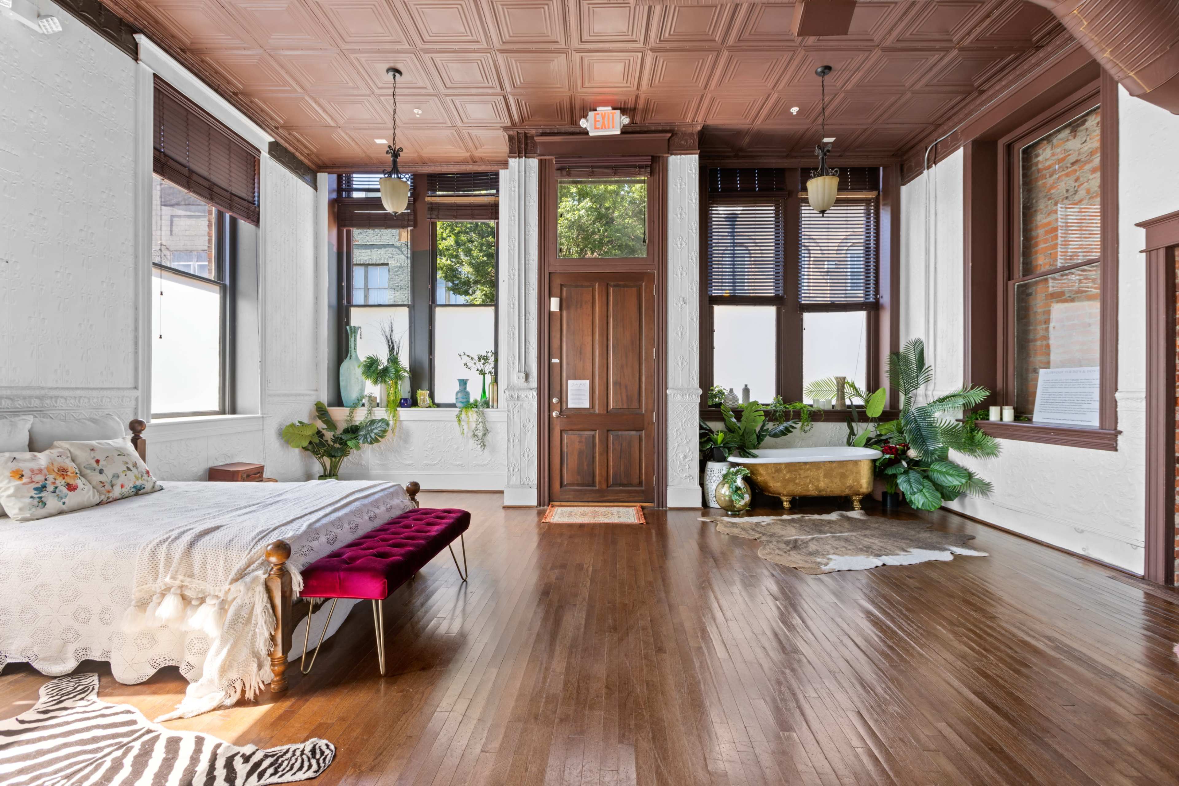 A spacious bedroom features a wooden bed with a pink bench, large windows, and plants decorating the area.