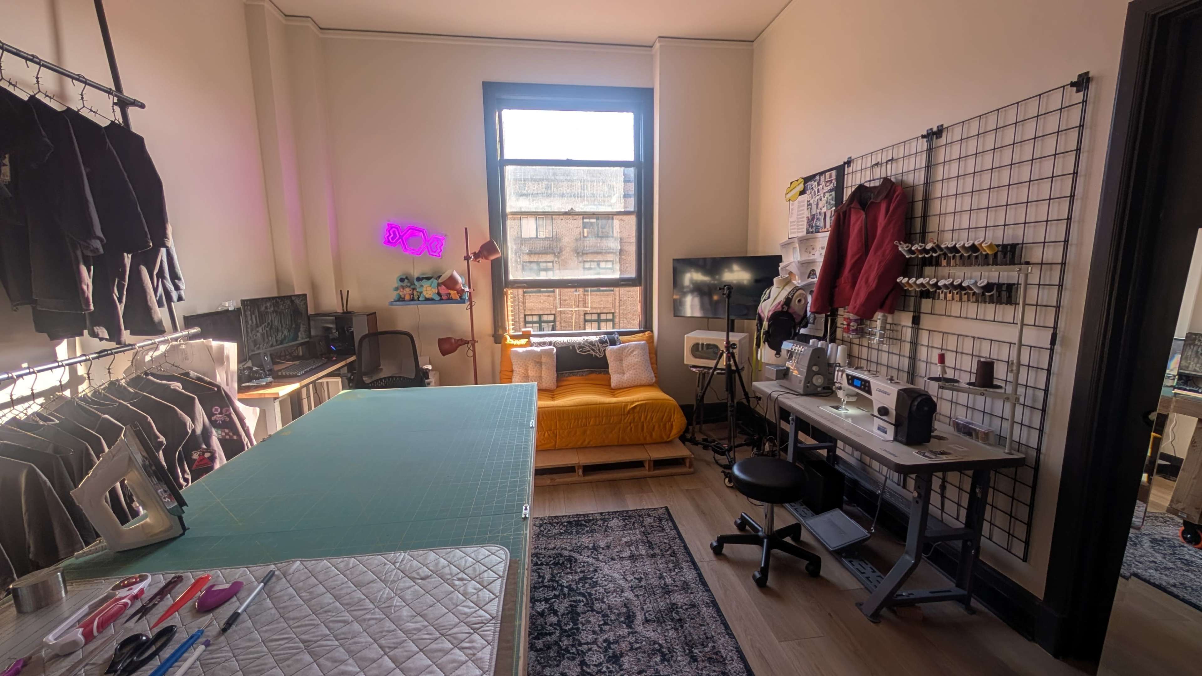 The image shows a well-organized workspace featuring a cutting table, sewing machine, and a yellow couch next to large windows with natural light streaming in.