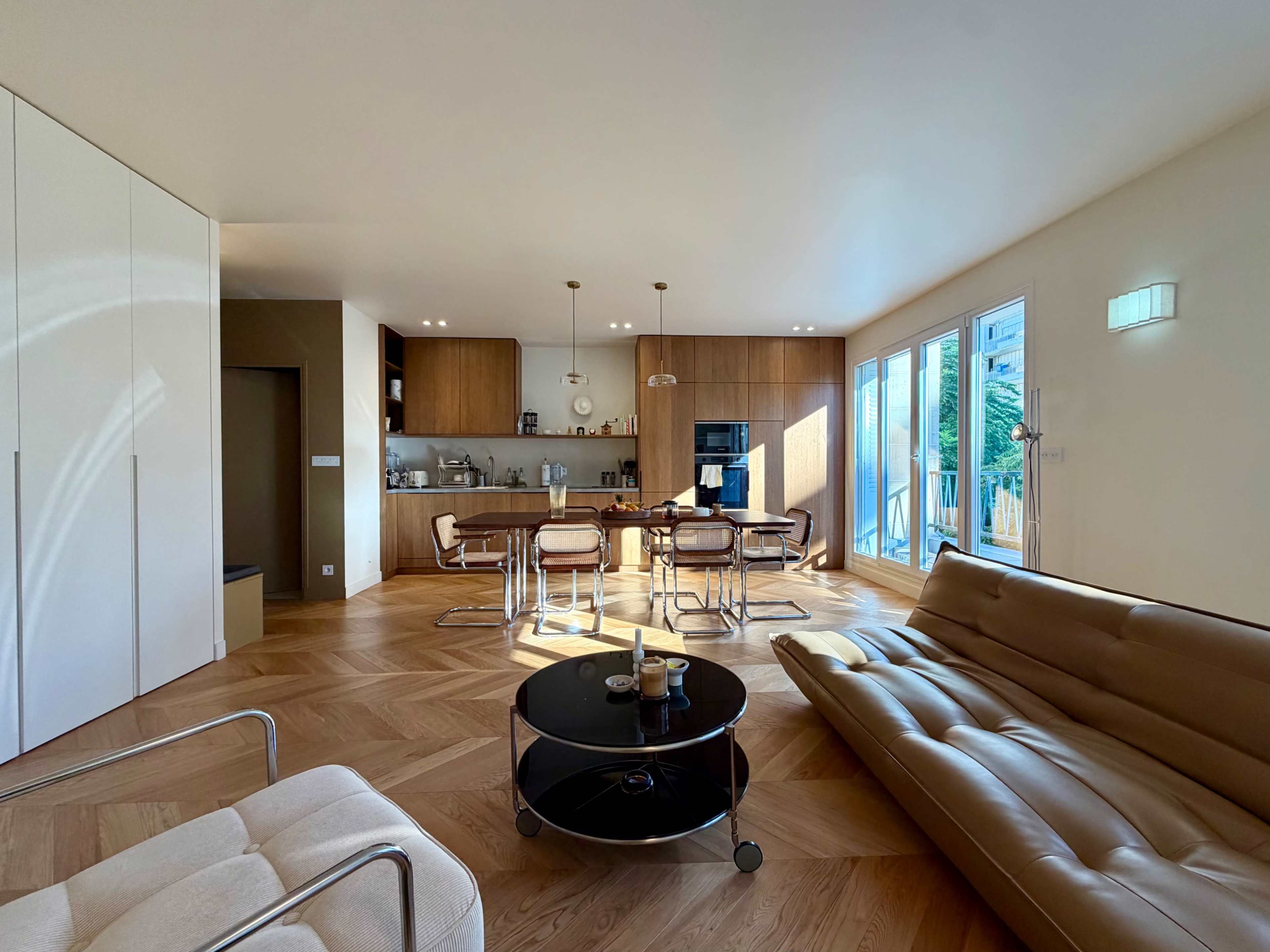 A modern living room with a kitchen area features a sofa, two chairs, and a round coffee table, illuminated by natural light from large windows.