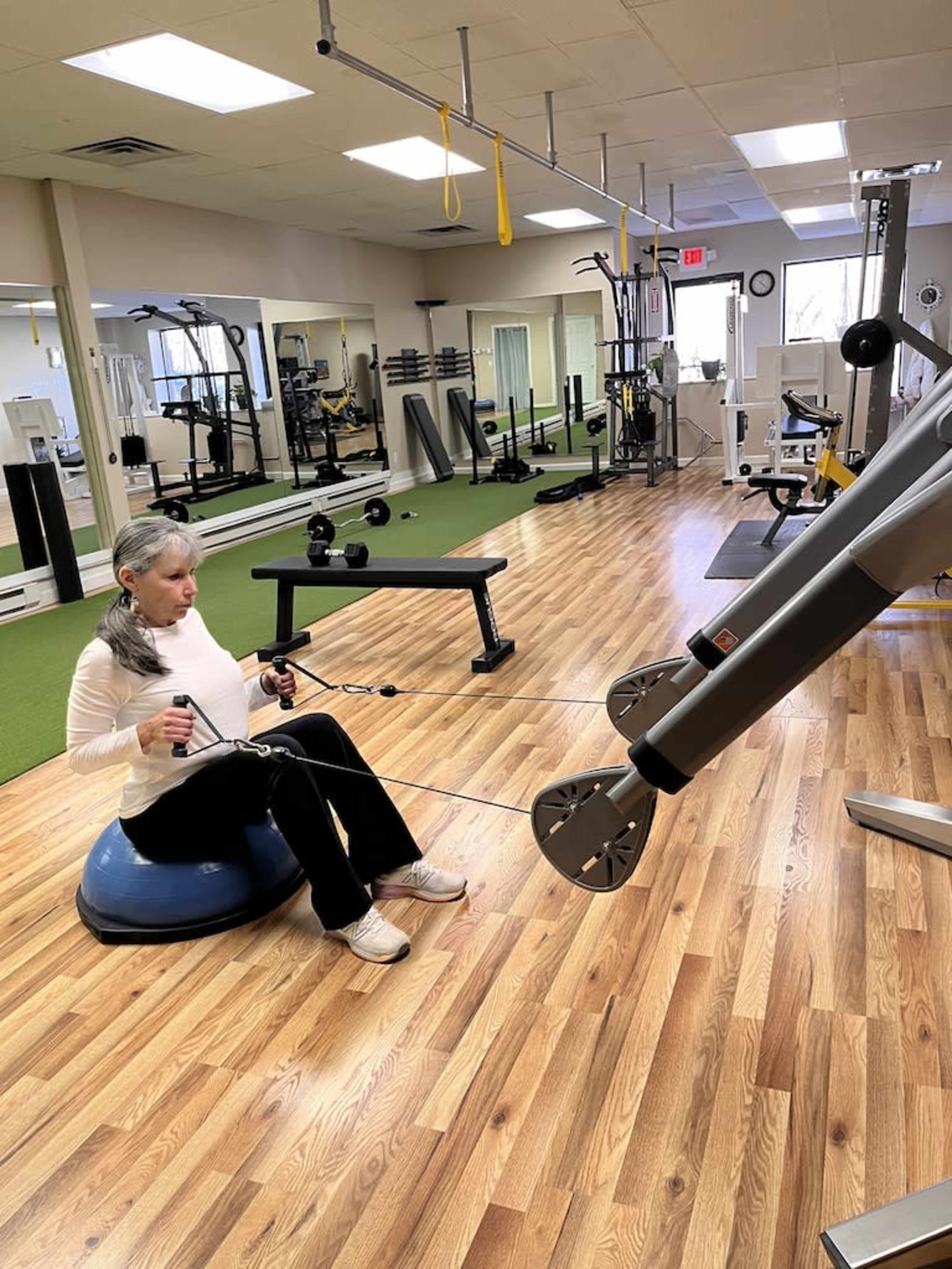A woman exercises on a stability ball while using a cable machine in a gym.