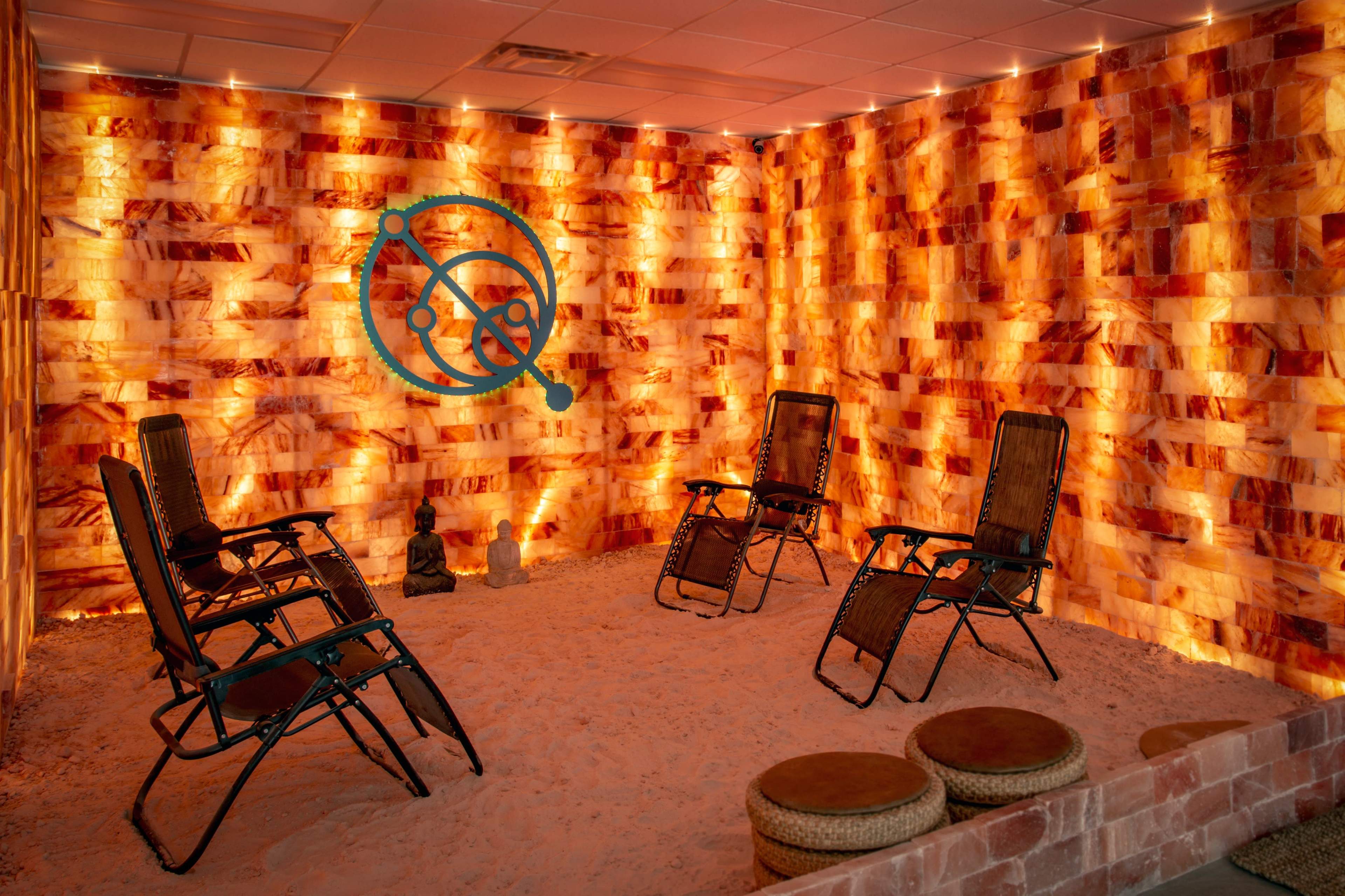 A therapy room with salt brick walls, illuminated by soft lighting, containing several chairs arranged in a circle on sandy flooring.
