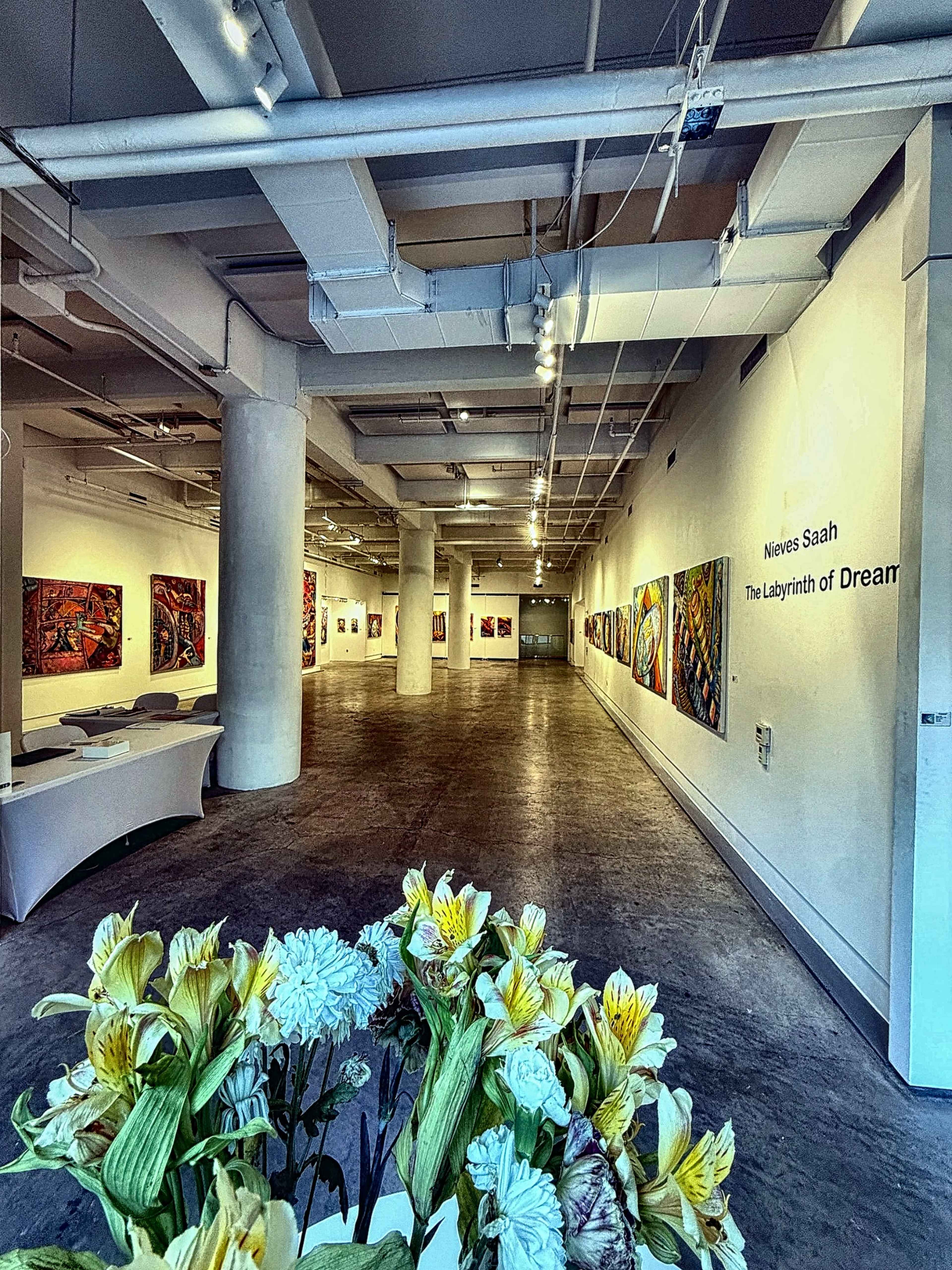 The image shows an art gallery with colorful paintings displayed on the walls, and a bouquet of flowers in the foreground.