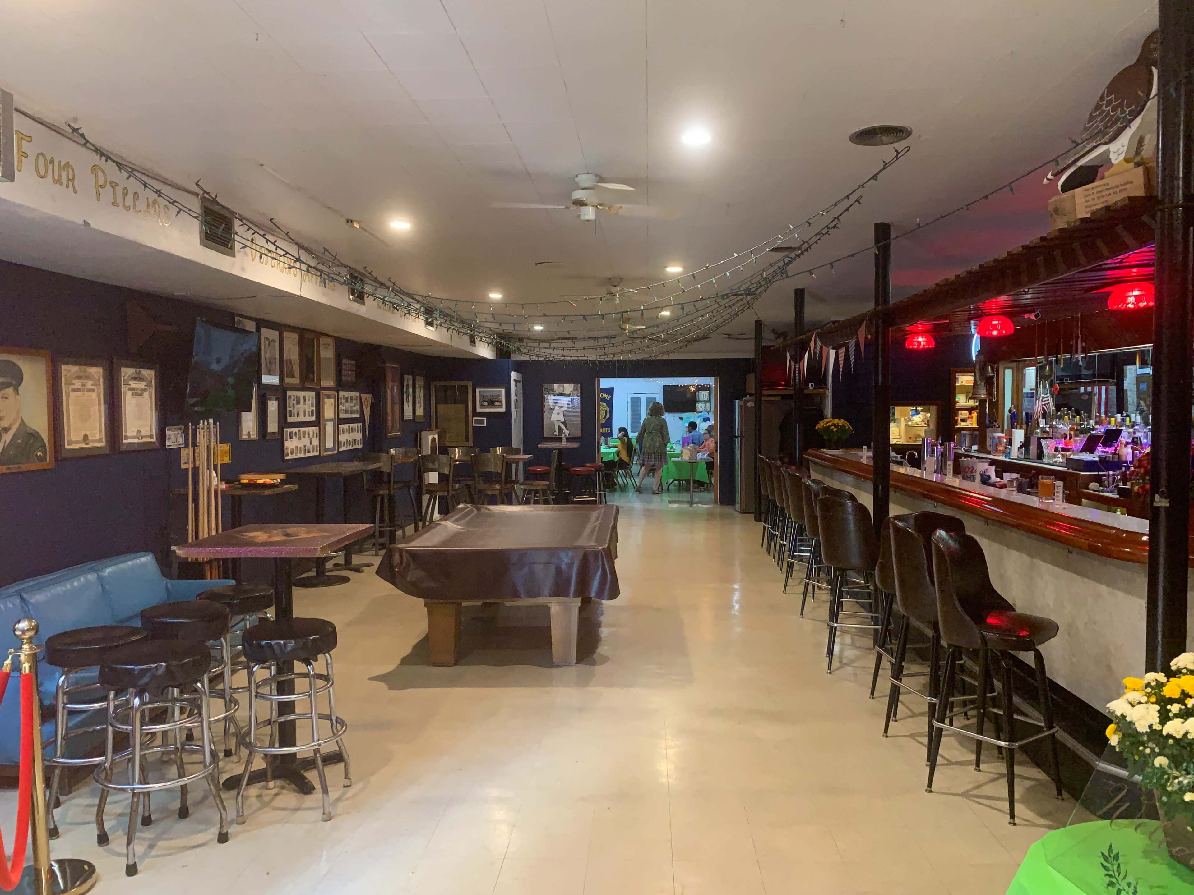 A dimly lit bar area with a pool table, seating at the bar, and a pathway leading to a room at the back with patrons.