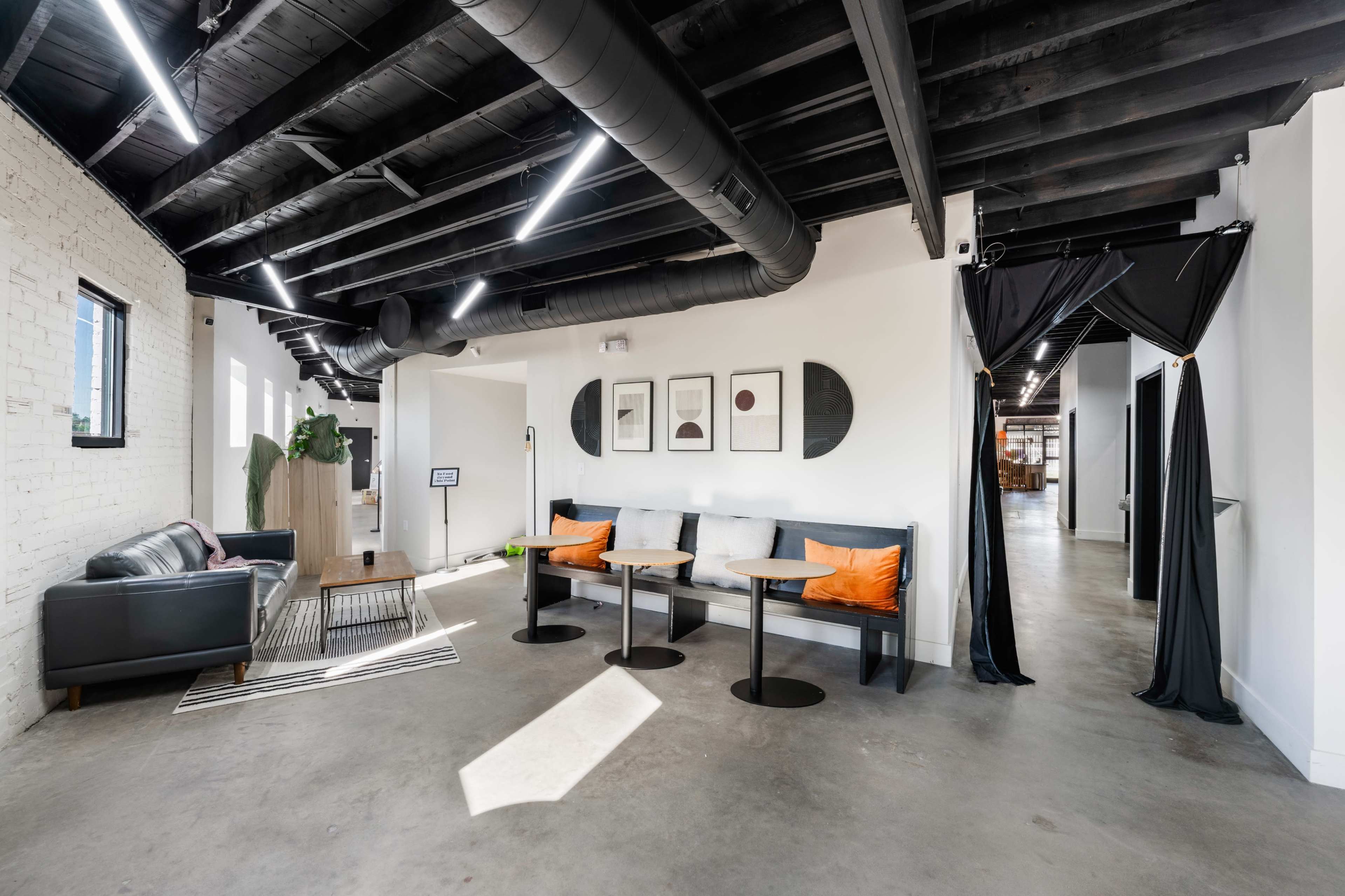 The image shows a modern office interior with a seating area featuring gray sofas and small black tables under a ceiling with exposed beams and ductwork.