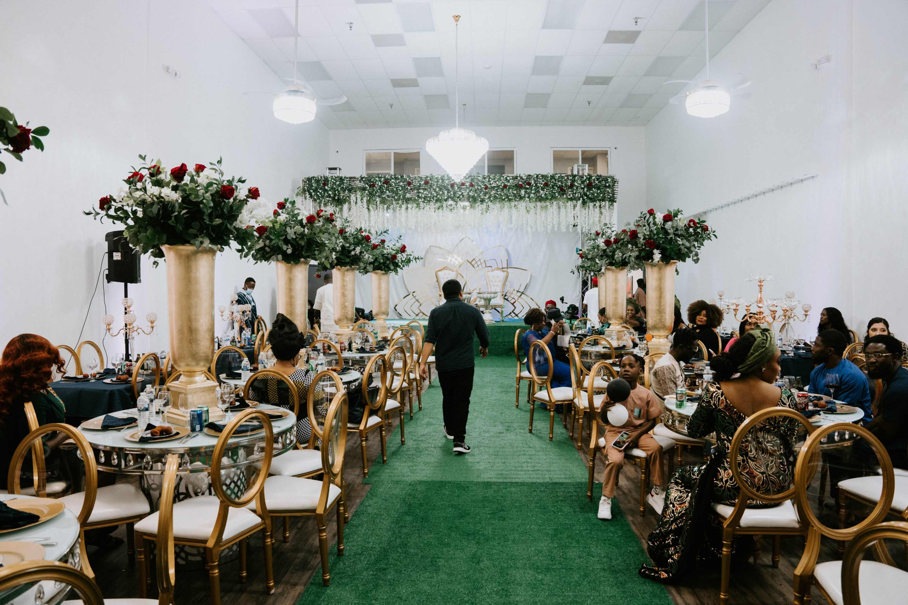 A banquet hall is set up for a formal event, featuring tables adorned with floral centerpieces, gold accents, and green carpet.