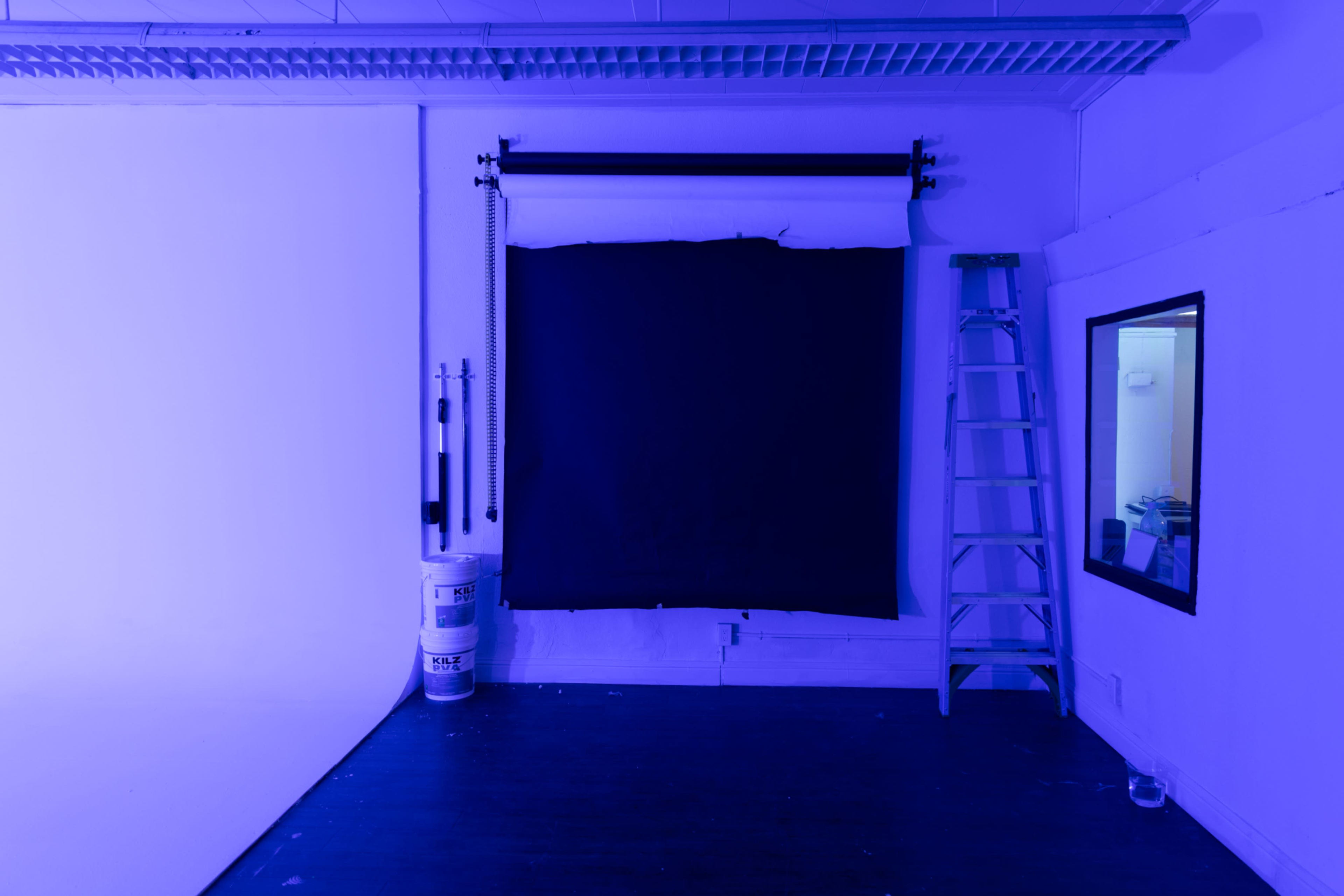 The image shows a photography studio with a purple backdrop, a white wall, a ladder, and a window reflecting the interior.