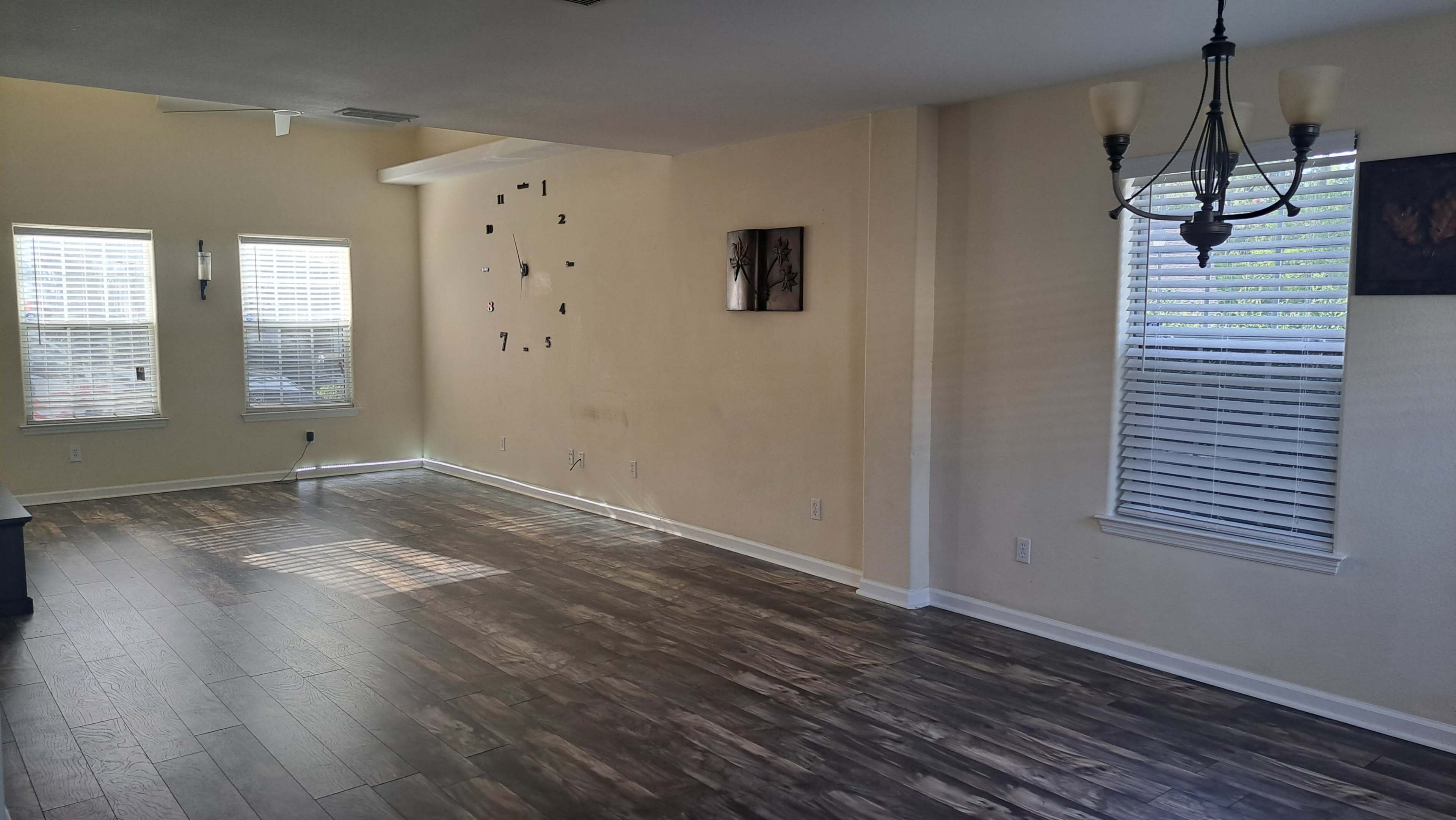 A well-lit room with wooden flooring, two windows featuring blinds, and a minimalist wall clock with numbers marked on the wall.