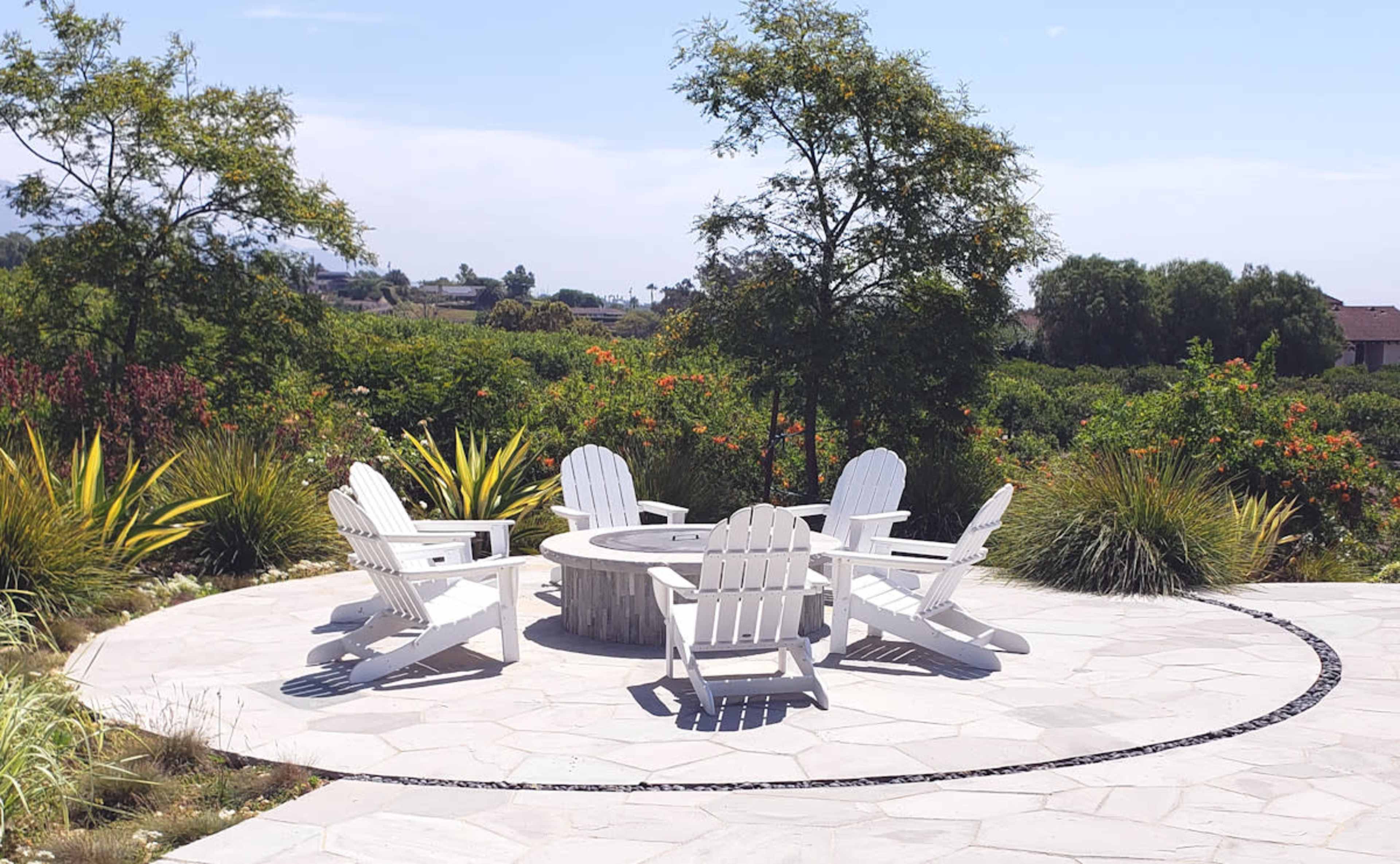 A circular stone patio features five white Adirondack chairs arranged around a fire pit, surrounded by greenery and trees.