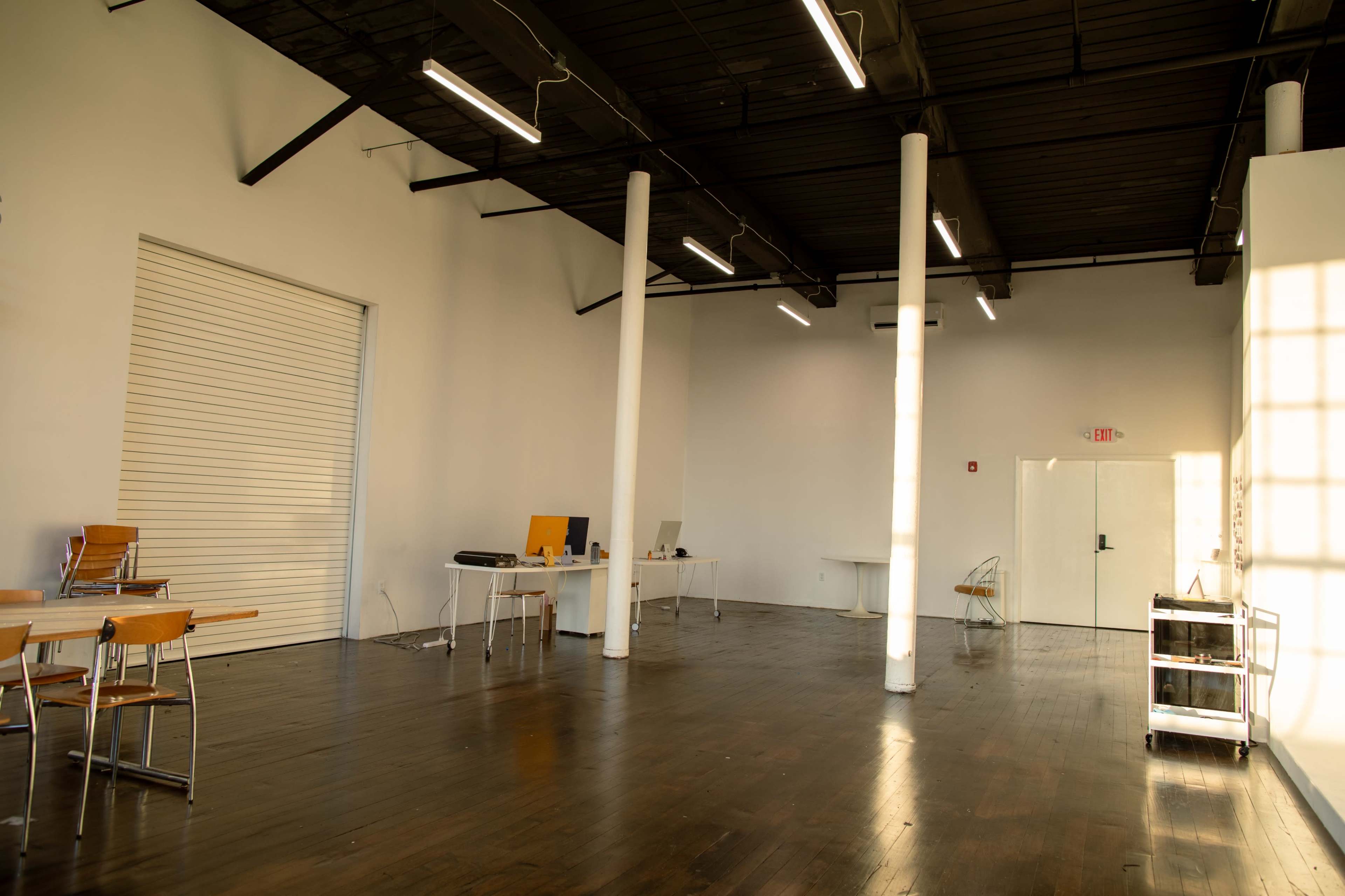 A spacious, empty interior of a modern office or studio with wooden floors and white walls, featuring several tables and chairs, along with a closed overhead door.