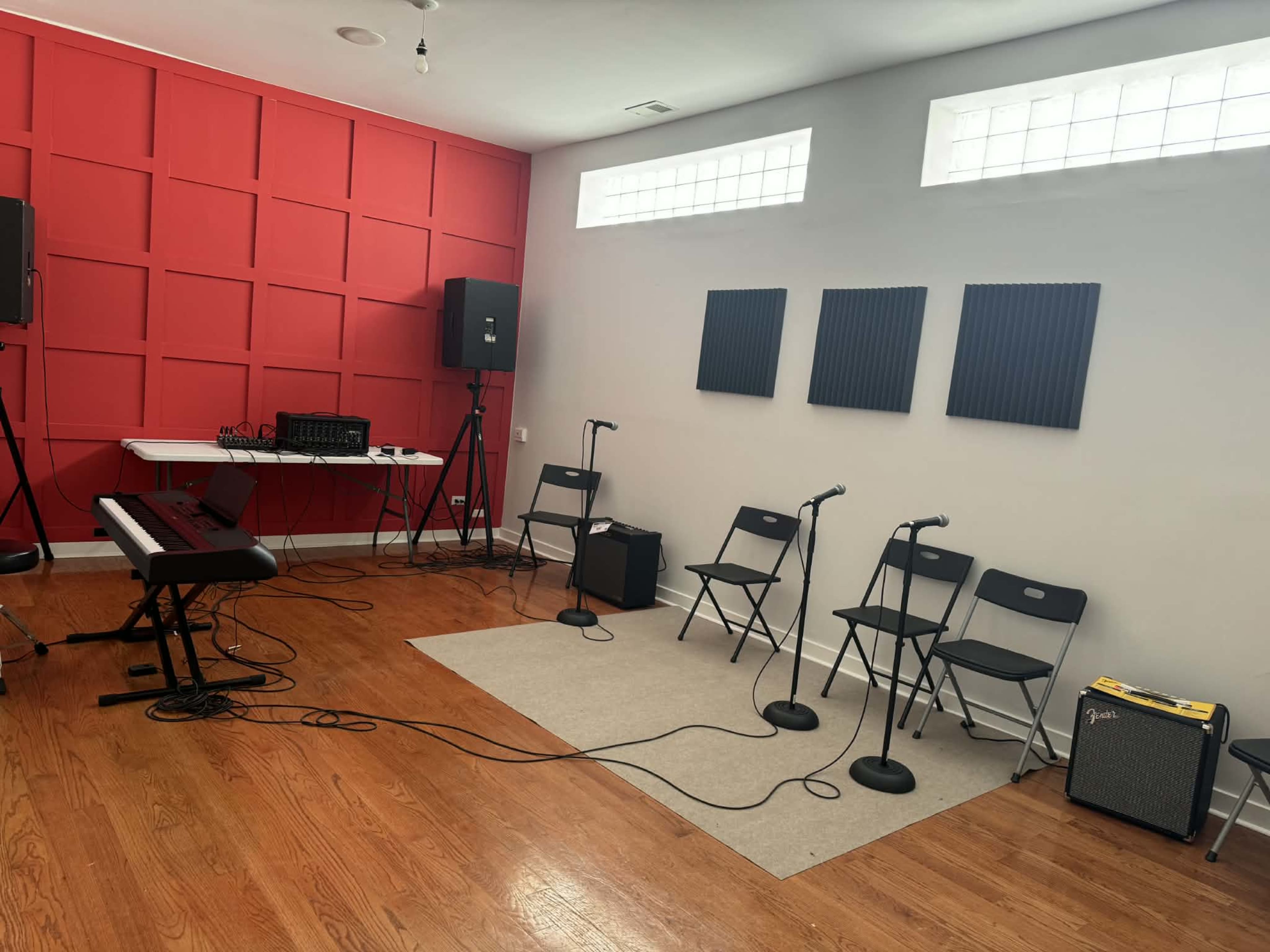The image shows a rehearsal room with a red accent wall, a keyboard, microphones on stands, several black chairs, and audio equipment.