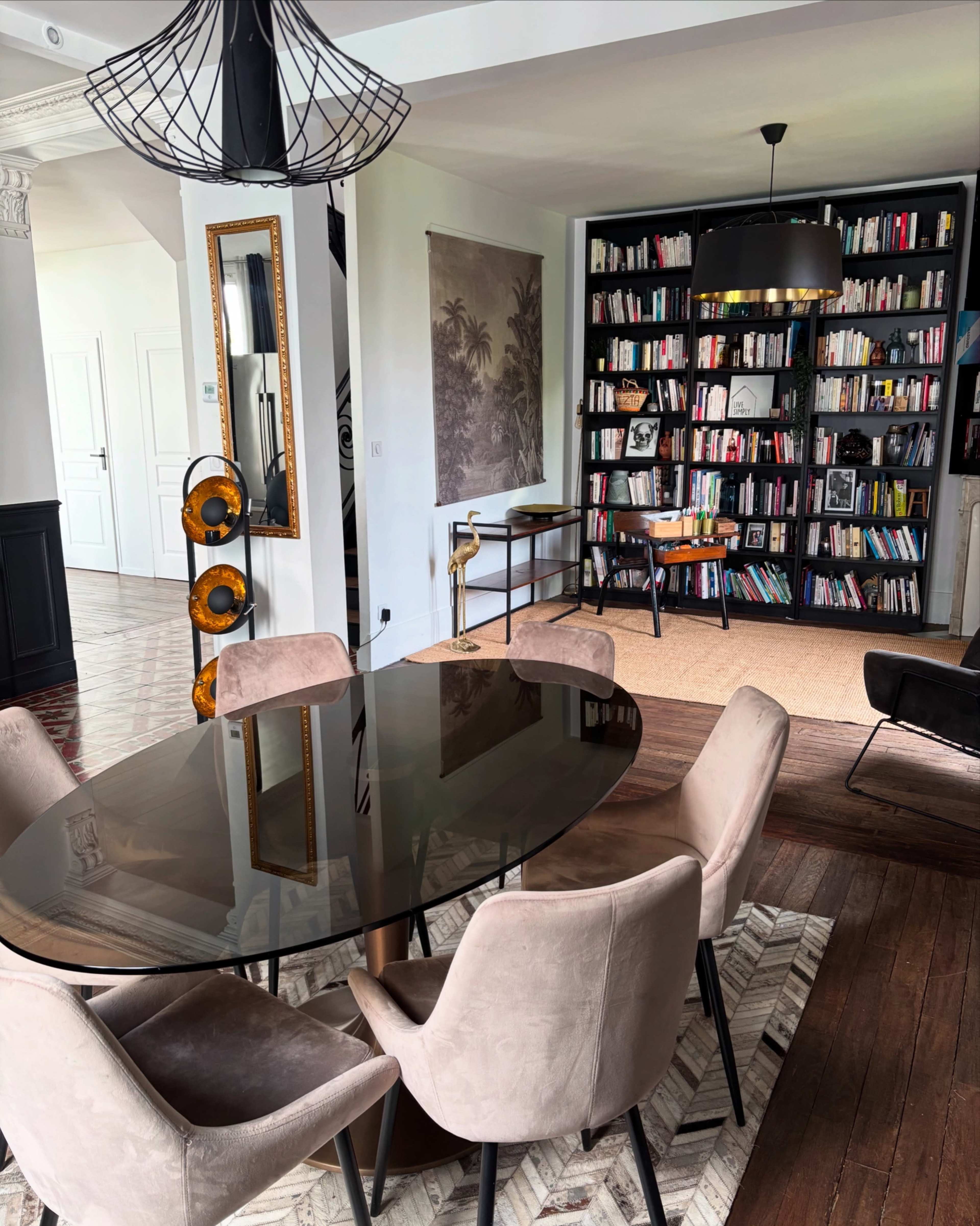 A dining area features an oval glass table surrounded by upholstered chairs, with a bookshelf and a chair visible in the adjacent room.