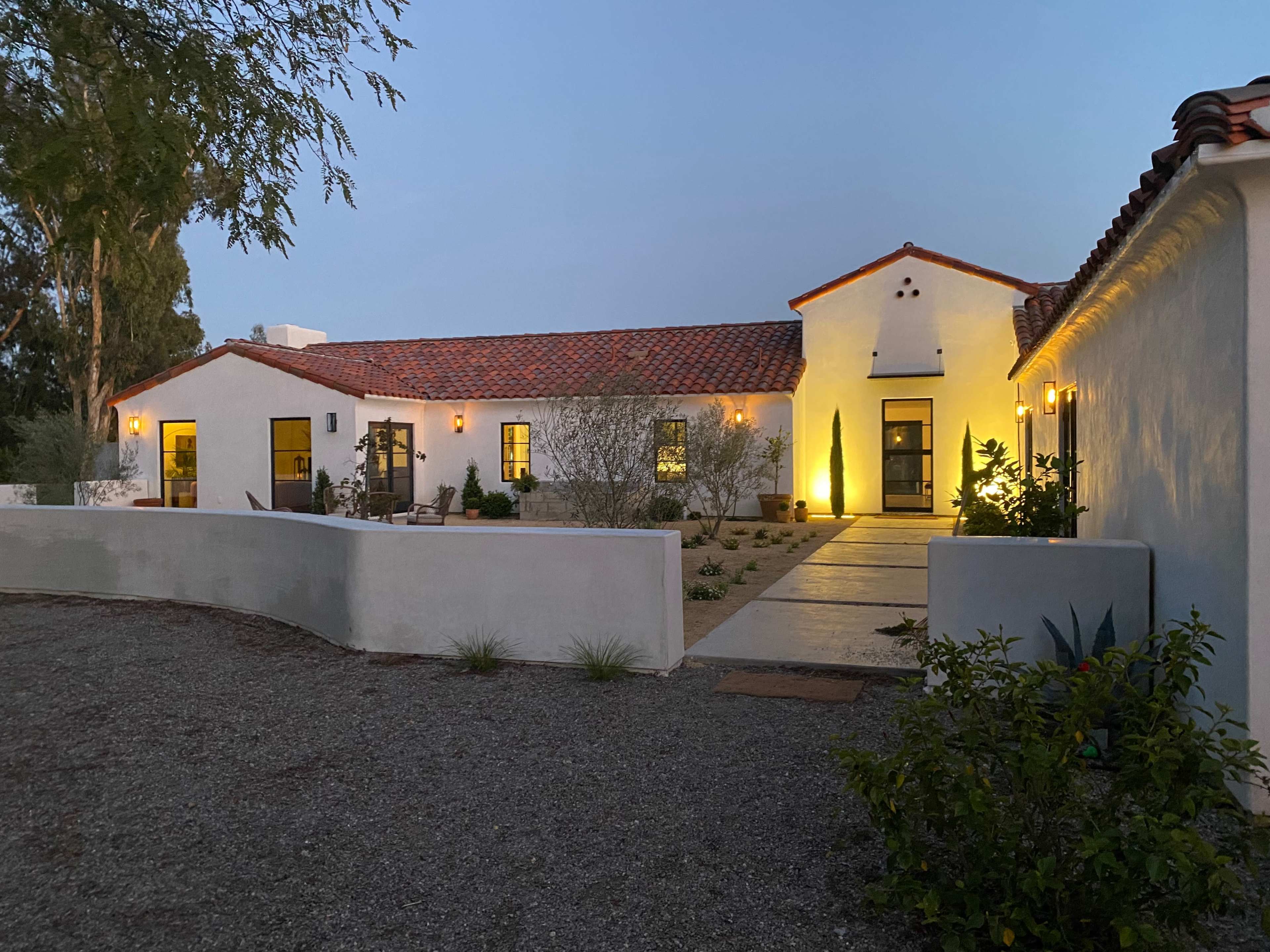 A modern single-story house with a red tile roof, white stucco walls, and landscaped gardens is illuminated in the evening light.