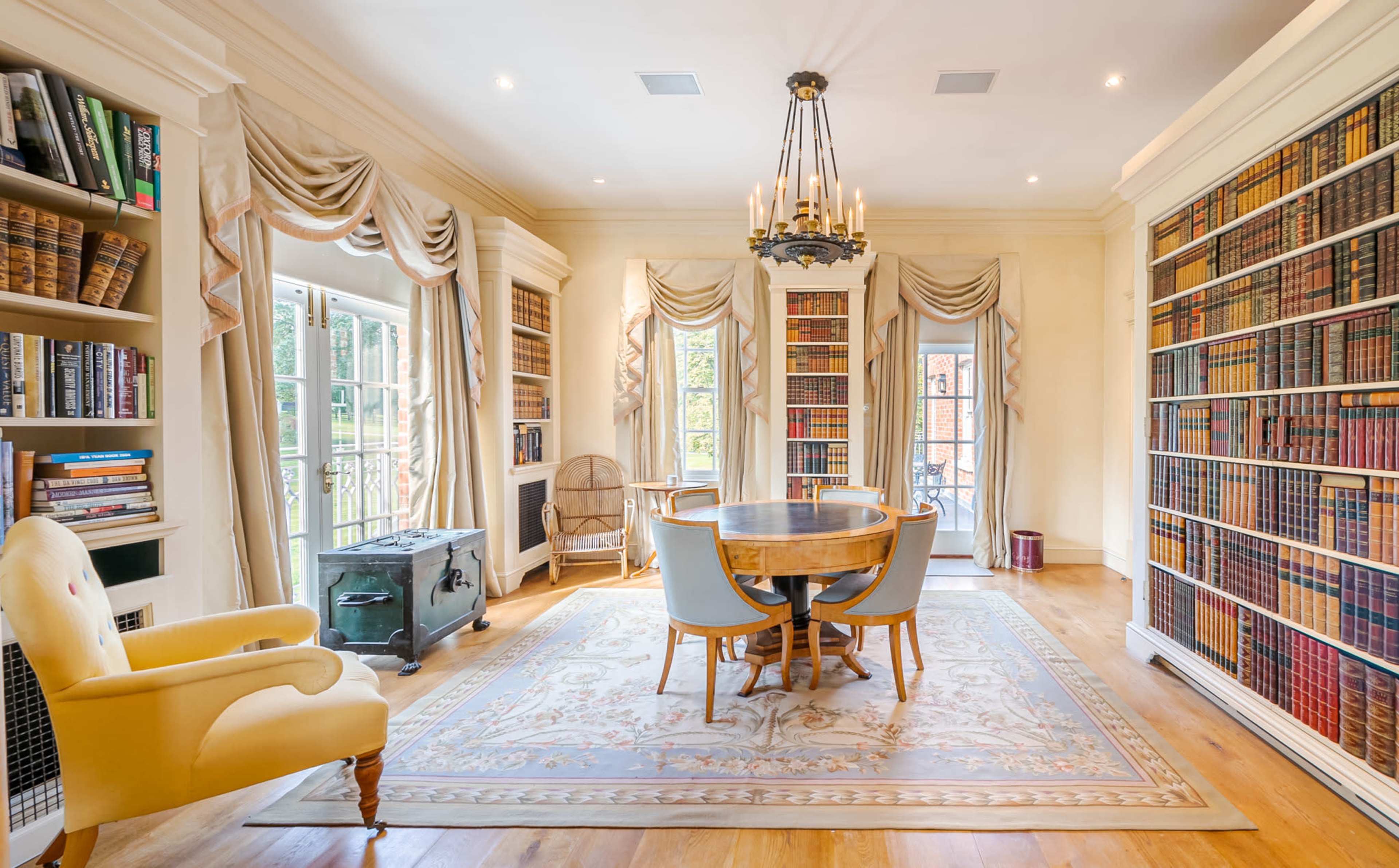 The image shows a well-lit library with floor-to-ceiling bookshelves, a circular table surrounded by blue chairs, and large windows with draped curtains.