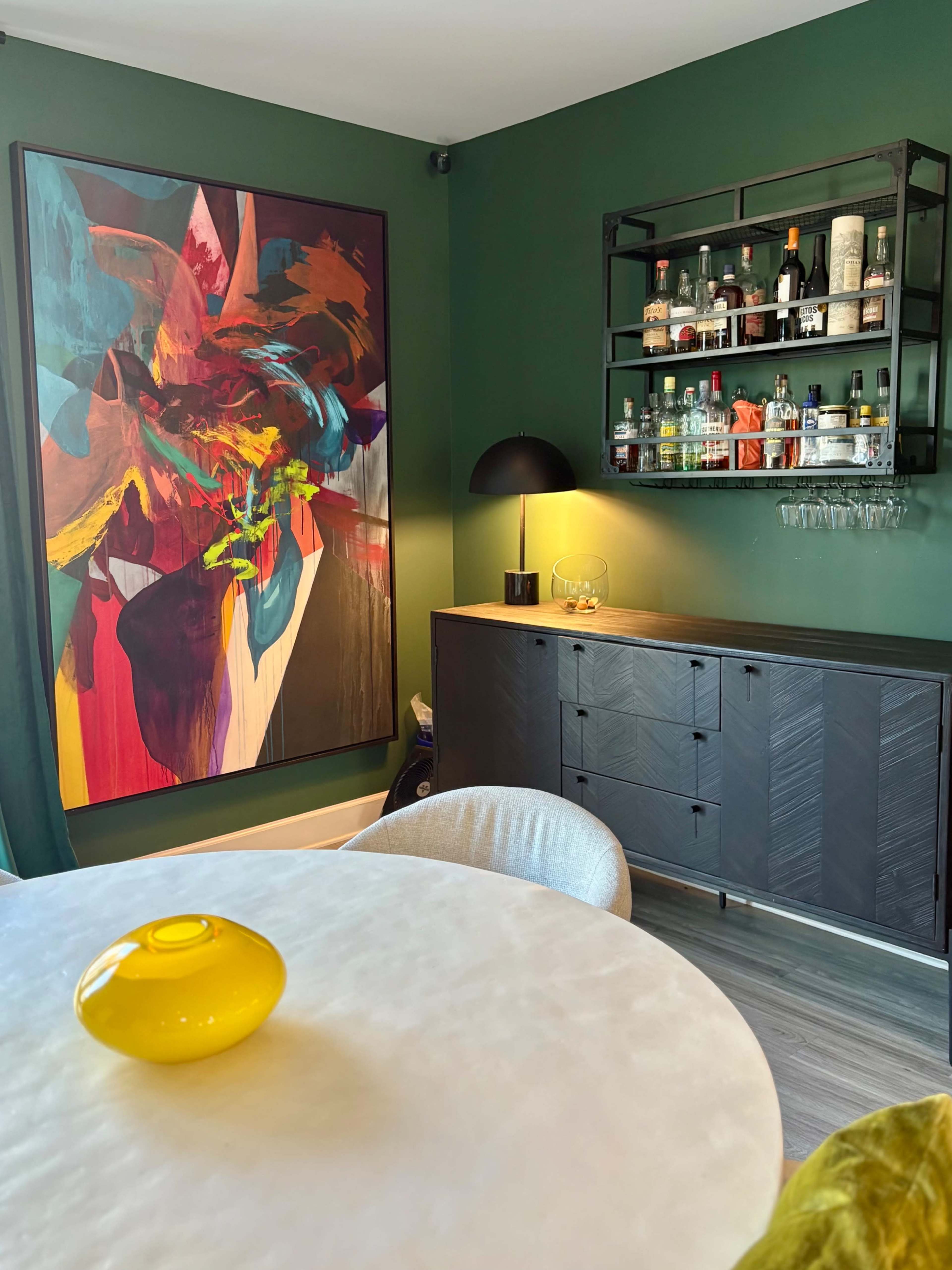 The image shows a modern living space with a bold abstract painting on a dark green wall, a round table with a yellow vase, and a black bar cabinet stocked with various bottles and glassware.