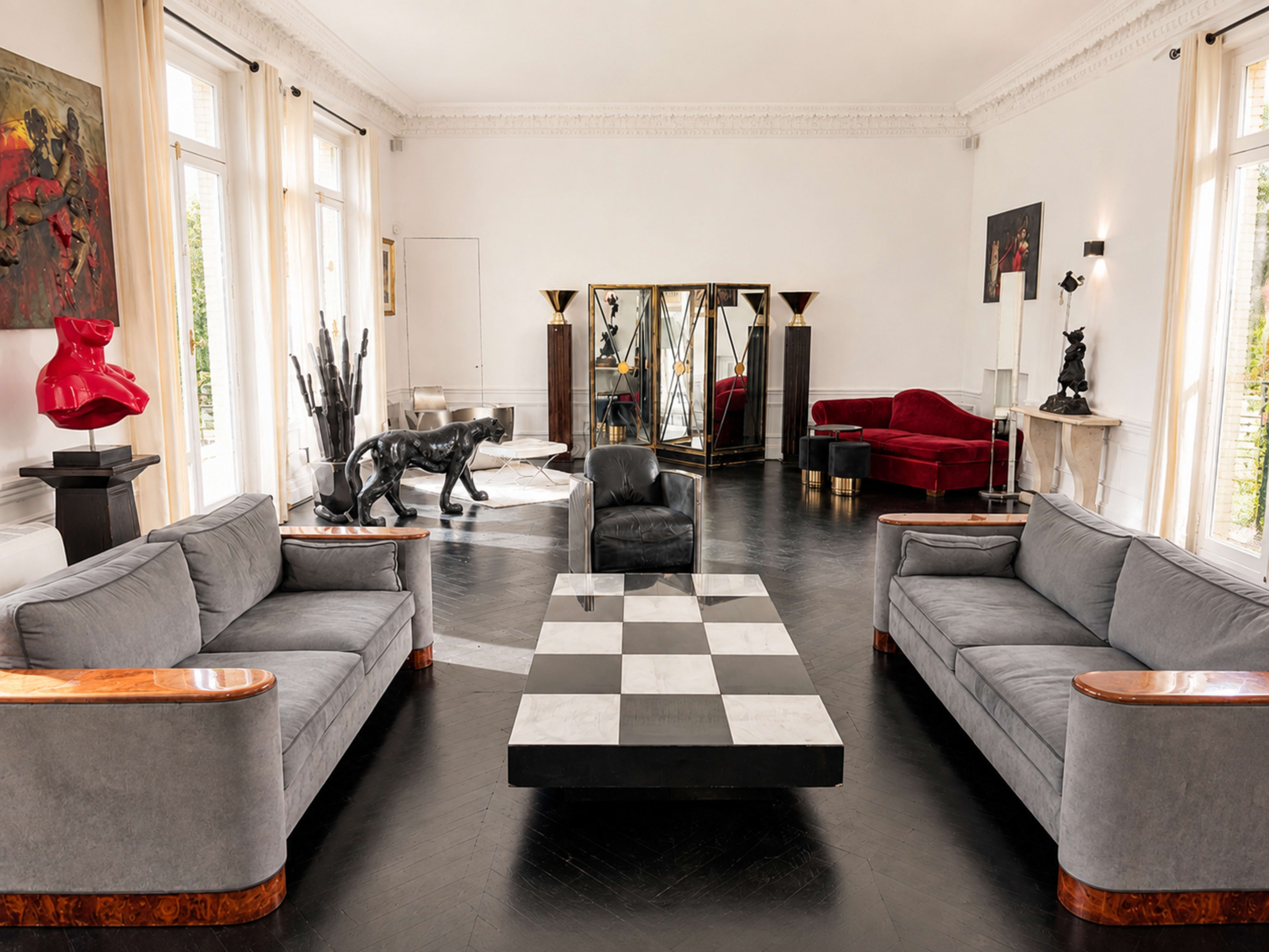 The image shows a modern living room with two gray sofas facing a large black and white checkered coffee table, a mirrored wall, and various artworks and sculptures.
