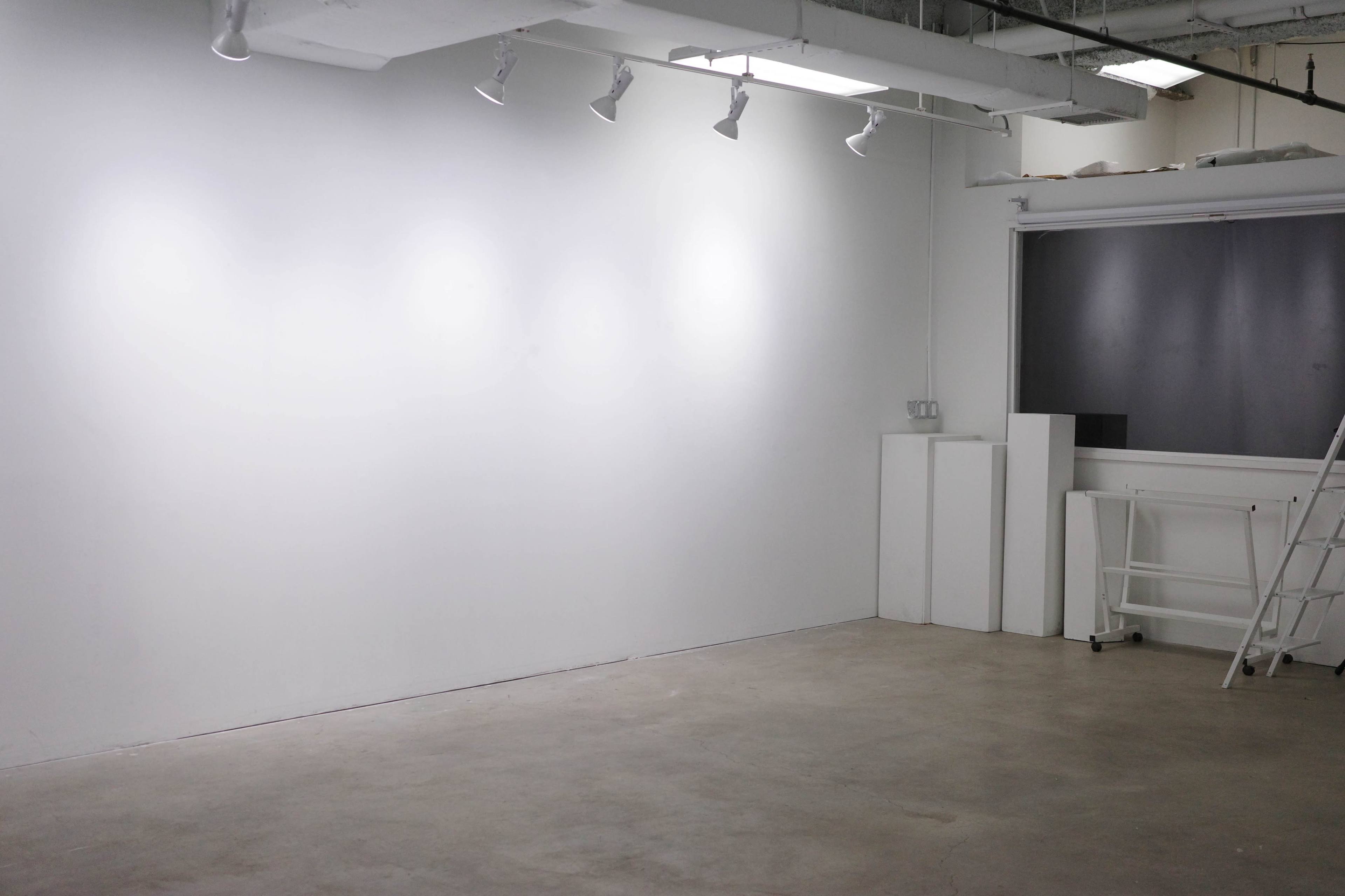 The image depicts an empty room with a white wall, overhead lights, and minimal furnishings, including a ladder and a small counter area.