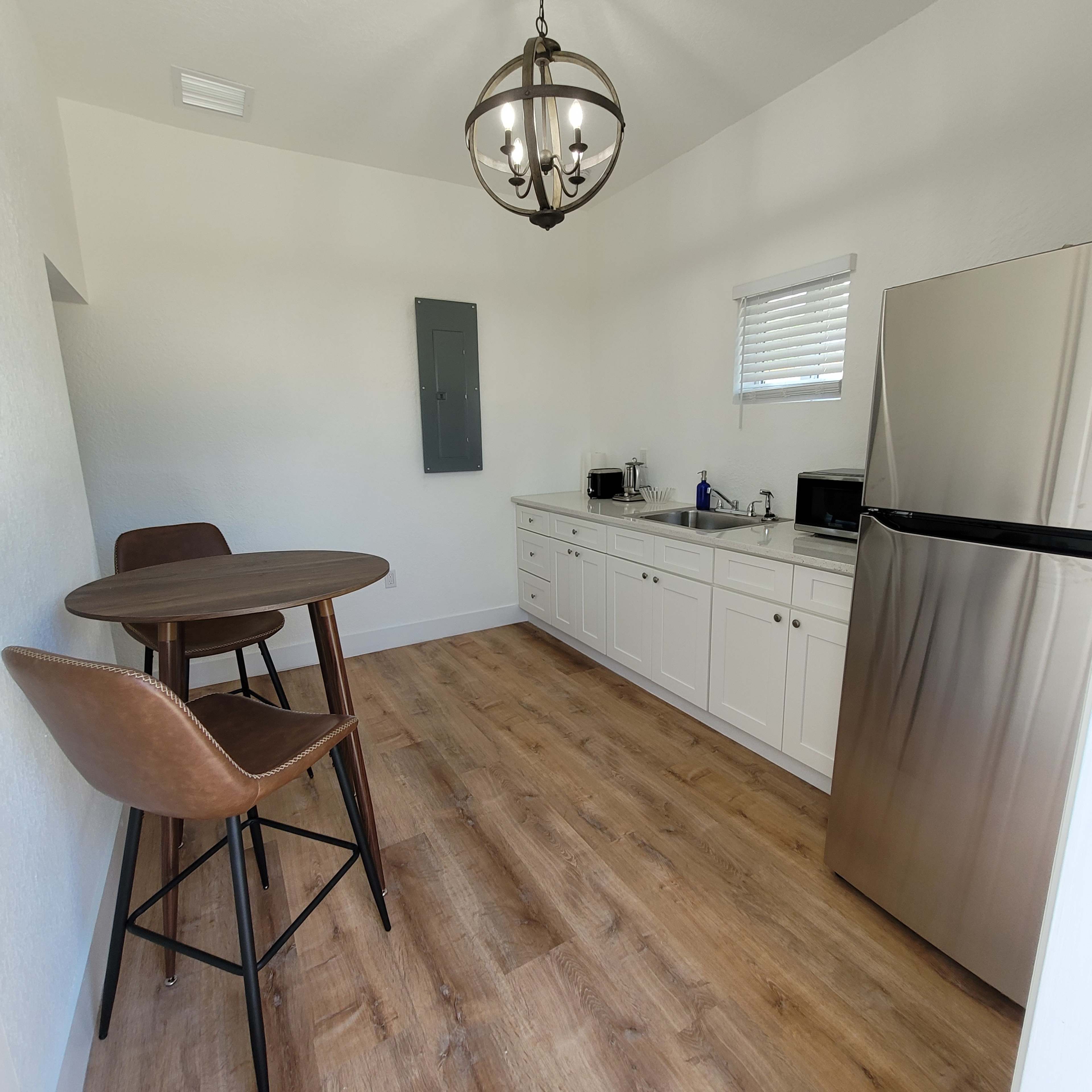 A small kitchen area features a round wooden table with two chairs, a stainless steel refrigerator, and white cabinetry with countertop appliances.