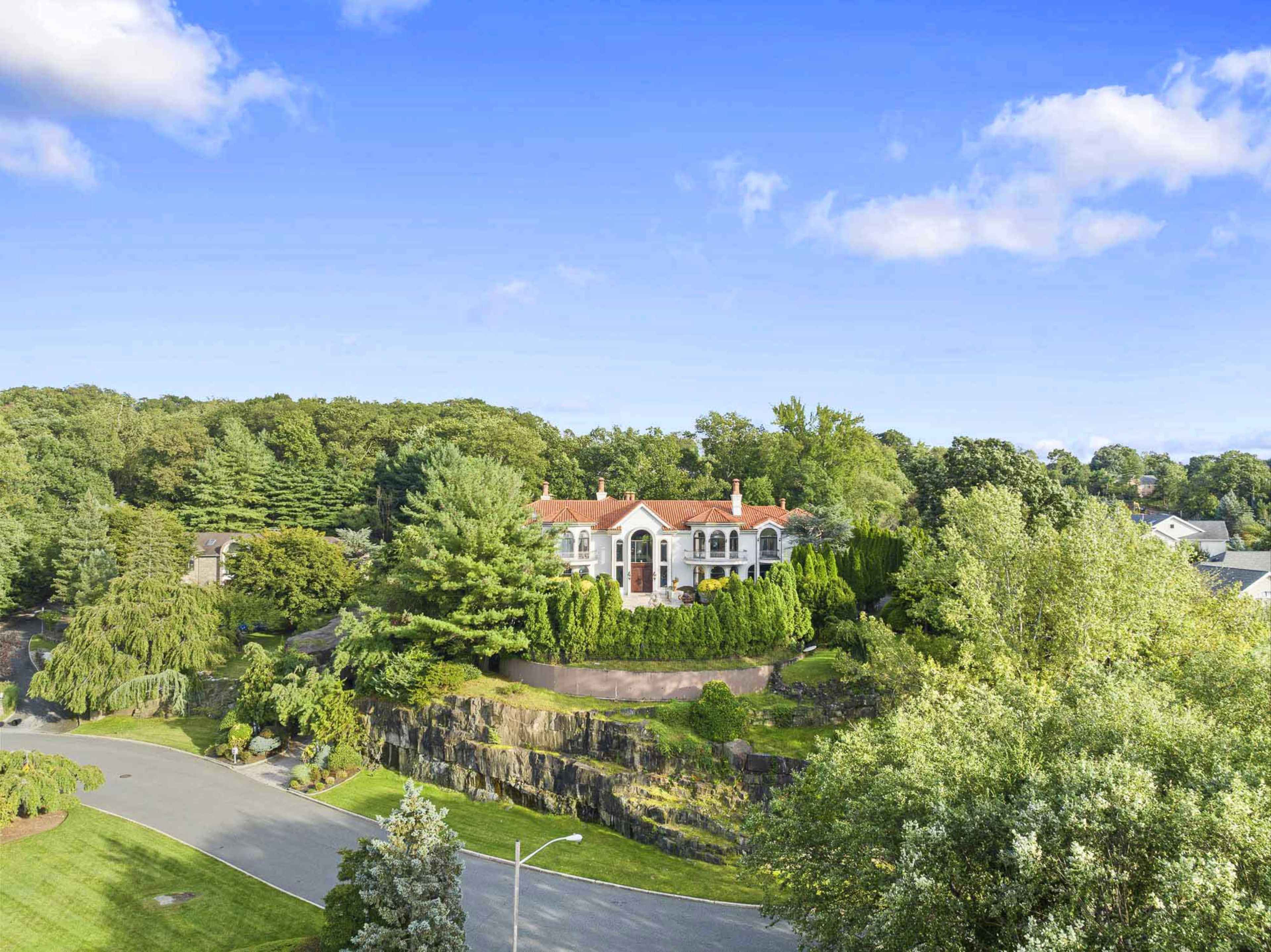 A large, two-story house with a red tile roof sits prominently on a hillside surrounded by trees and greenery.