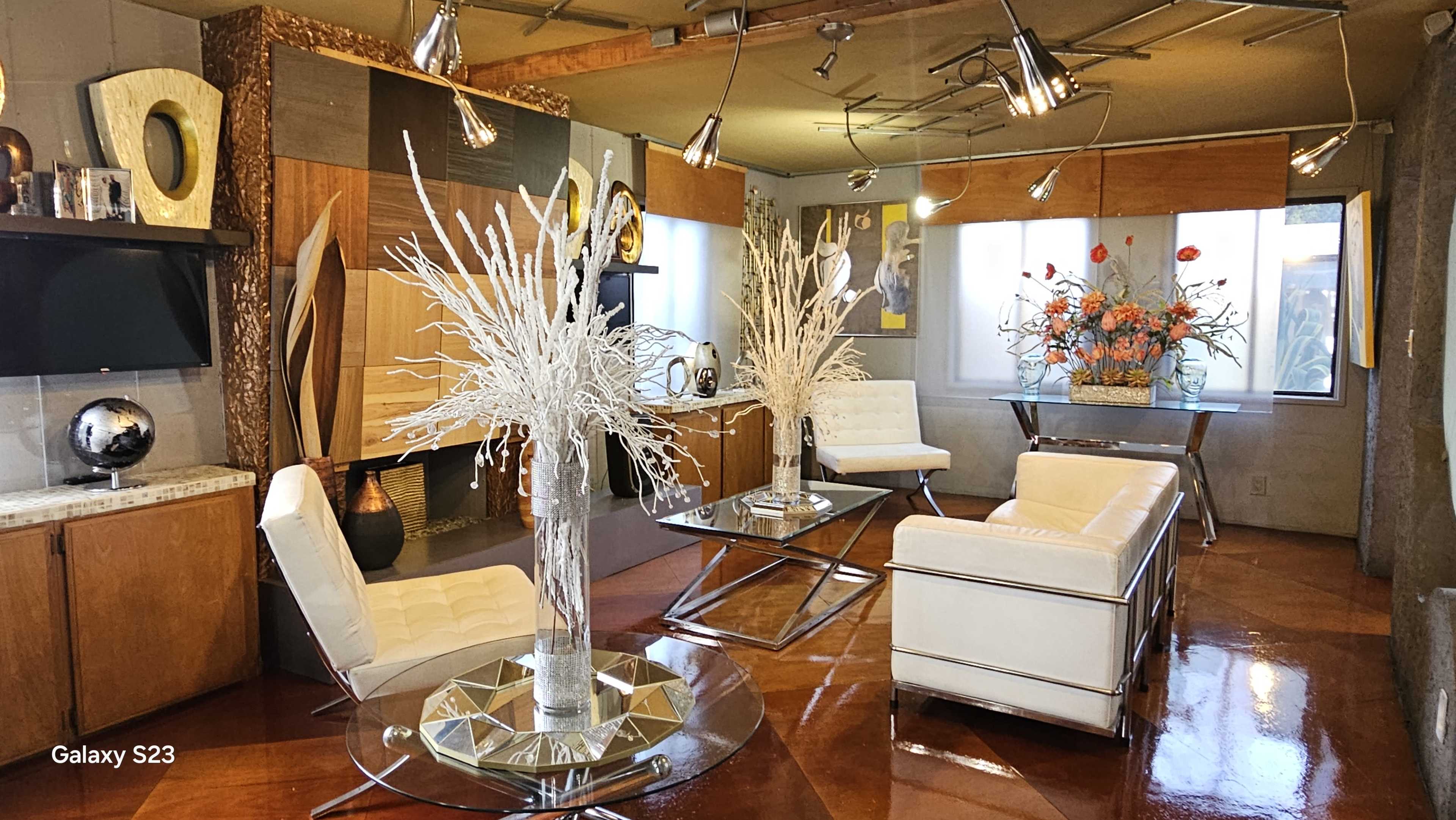 The image shows a modern living room with white furniture, decorative vases, and a large floral arrangement on a table.
