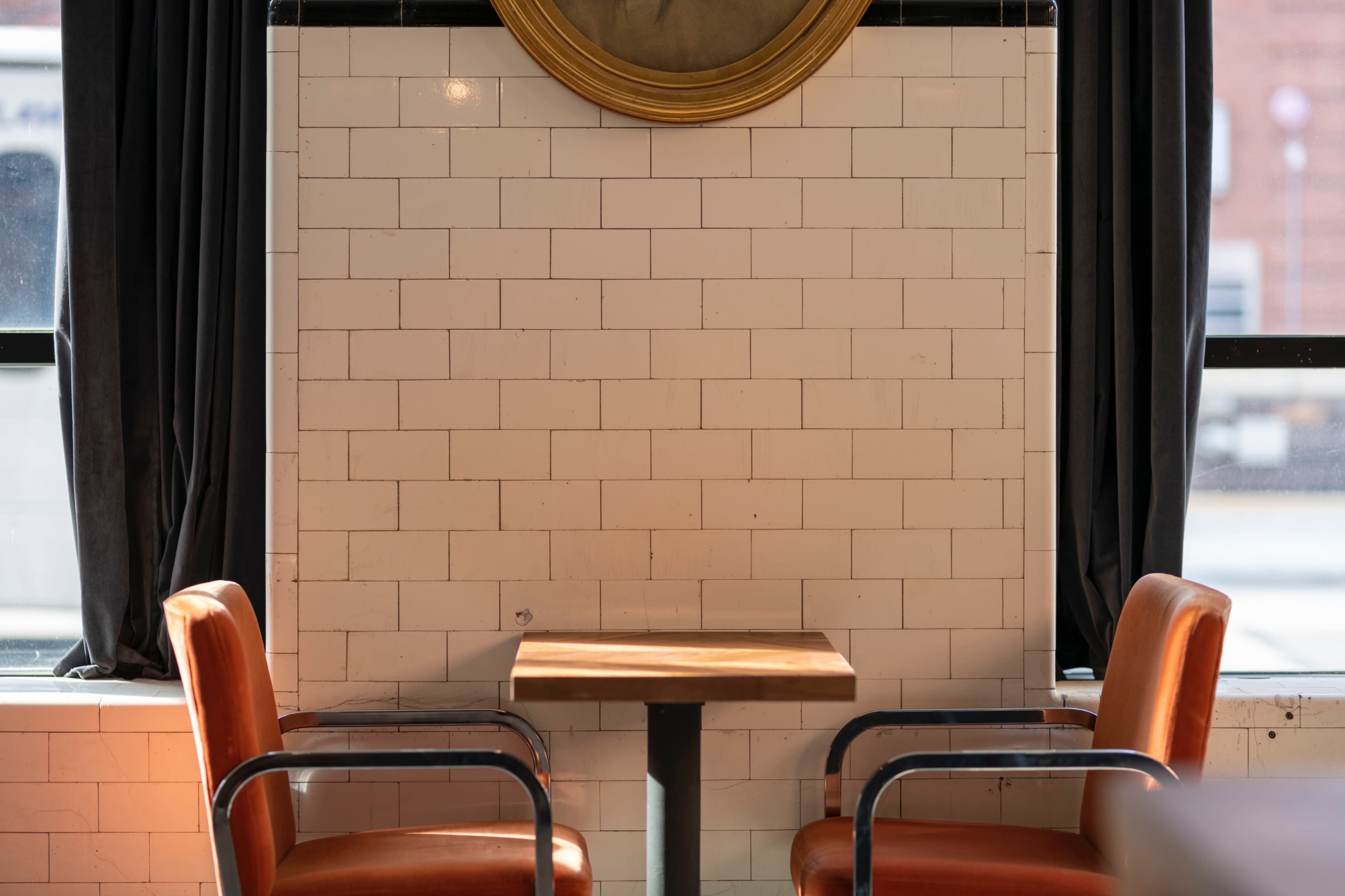 A small wooden table is positioned between two brown leather chairs in front of a tiled wall beneath a circular framed mirror.