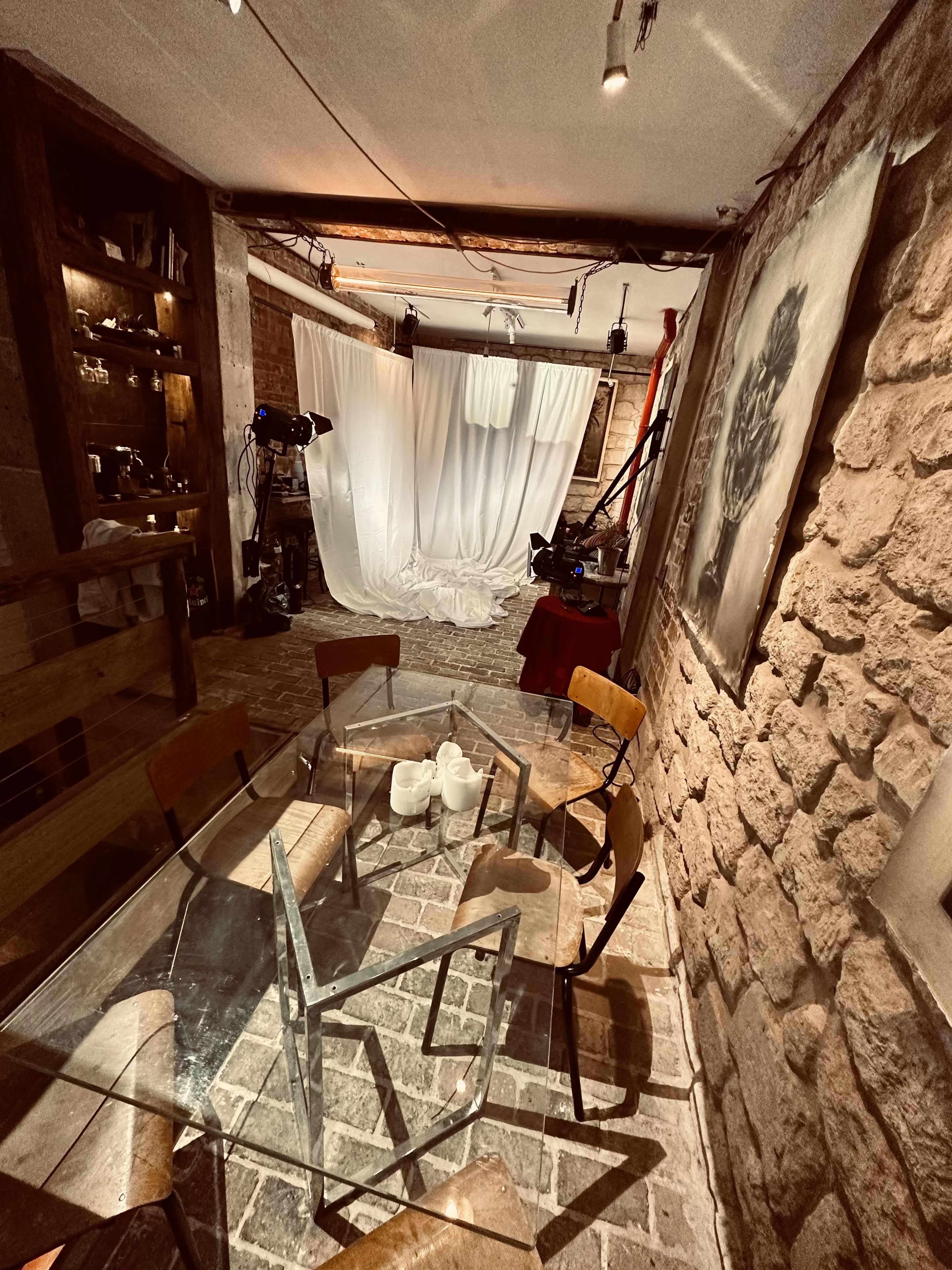 A room with a stone wall, a glass dining table surrounded by chairs, and a backdrop of white fabric behind a camera setup.