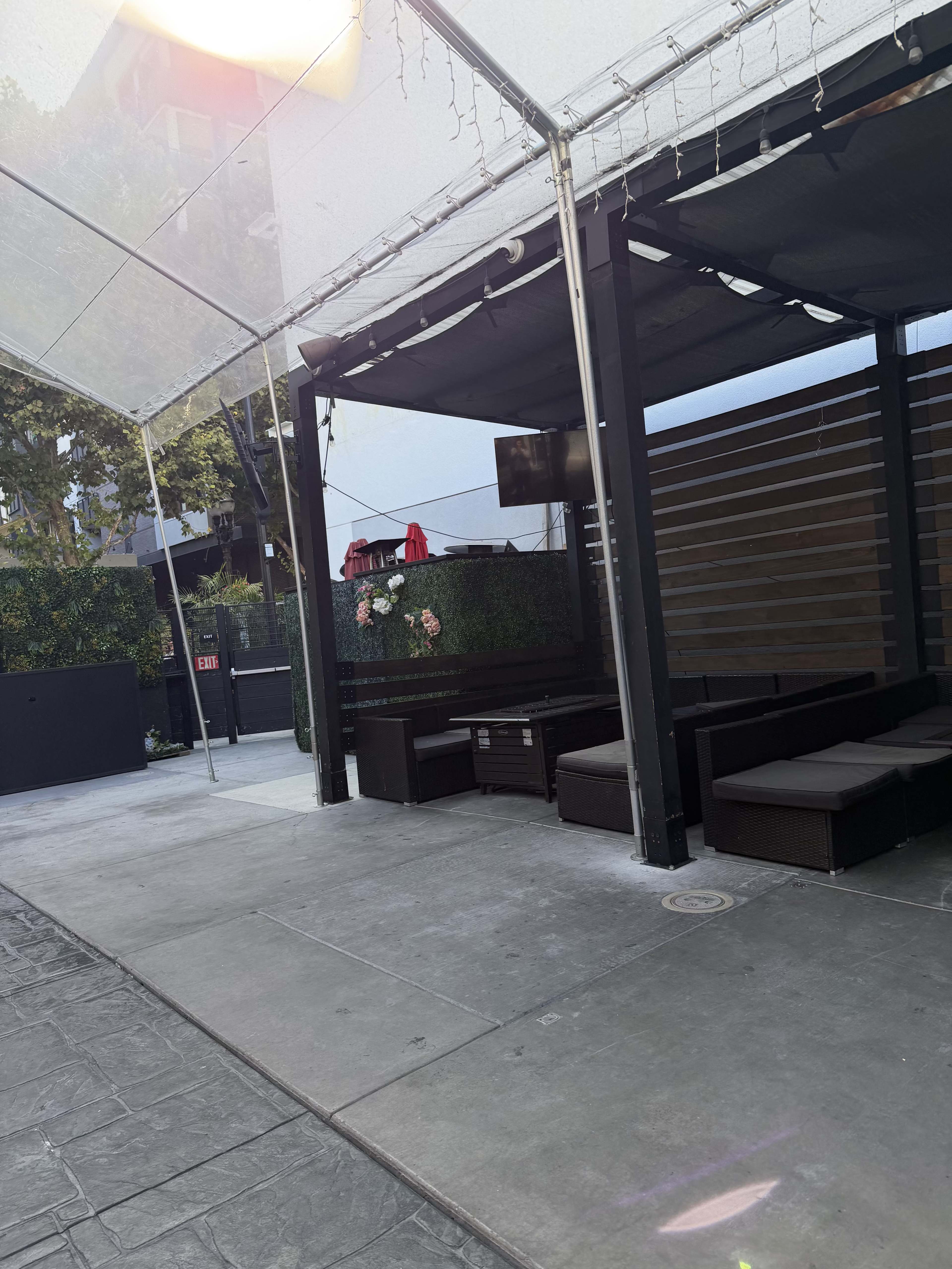 The image shows an outdoor seating area of a patio covered by a tent, featuring dark furniture and plants along the walls.