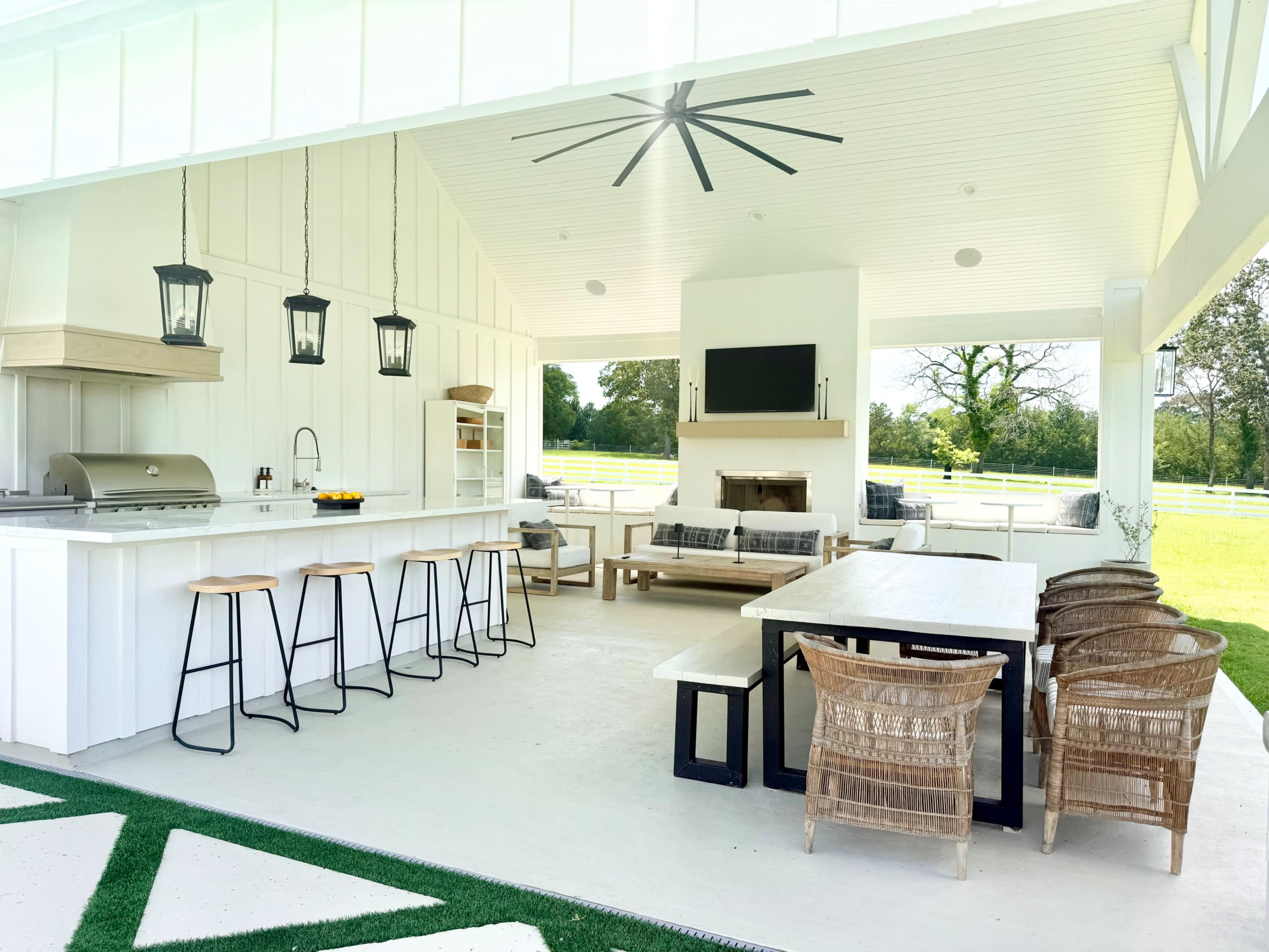 The image shows a modern outdoor living space featuring a kitchen area with a grill, a dining table, and seating arrangements, all set under a roof with a fan.
