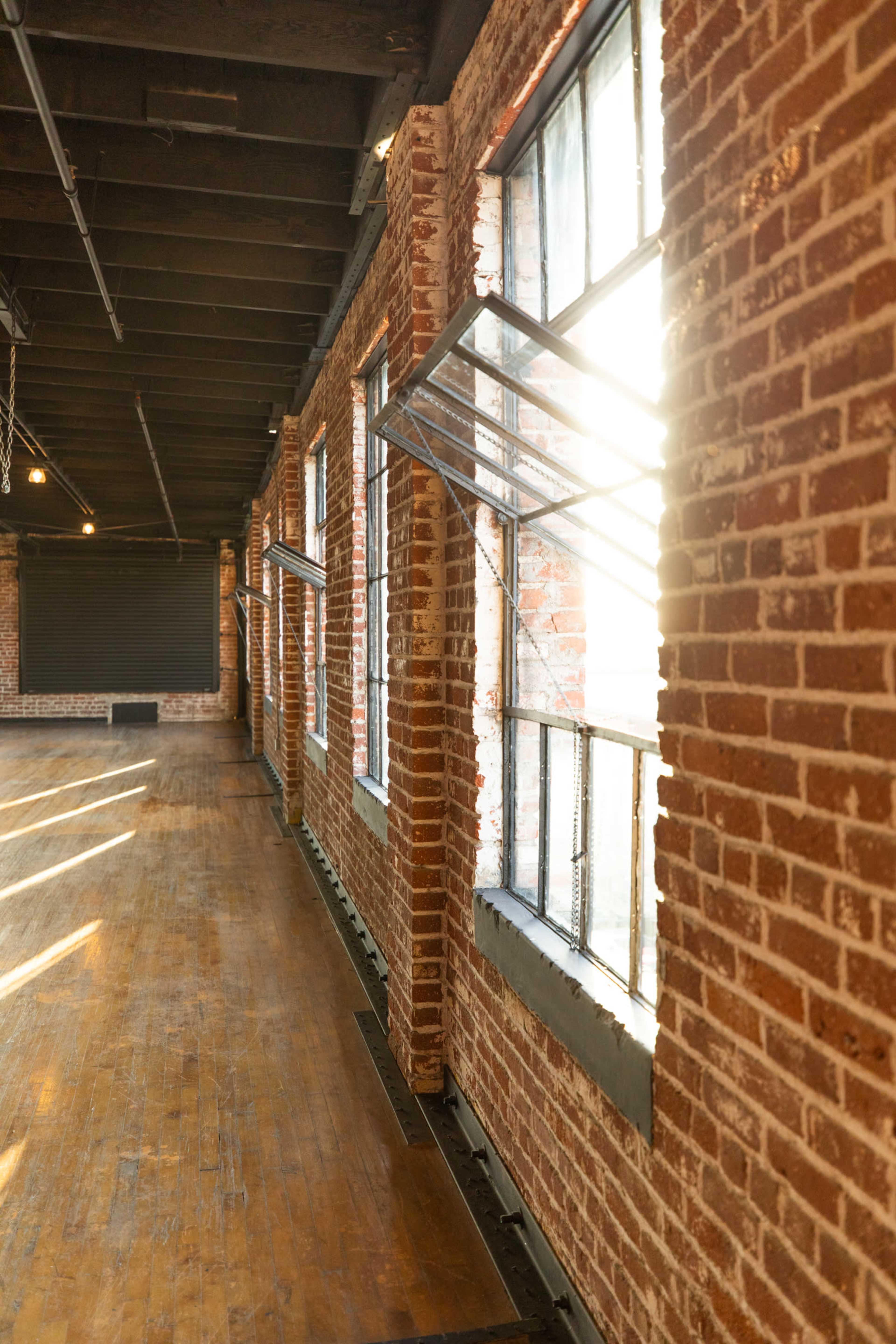 A spacious room with exposed brick walls, large windows, and wooden flooring, illuminated by sunlight streaming through the windows.