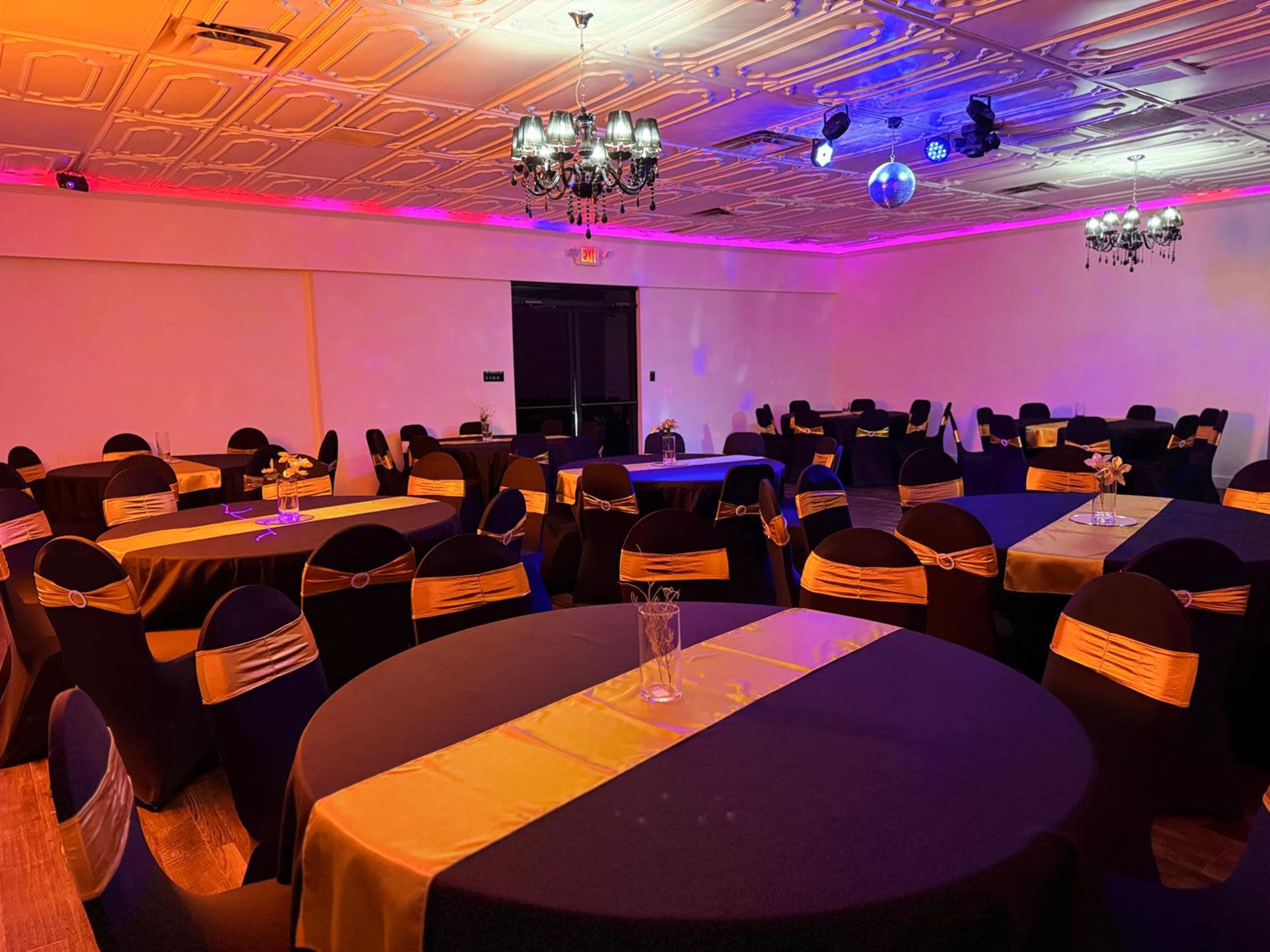 The image shows a banquet hall set up with round tables covered in black and gold tablecloths, surrounded by chairs adorned with gold sashes under colorful lighting.