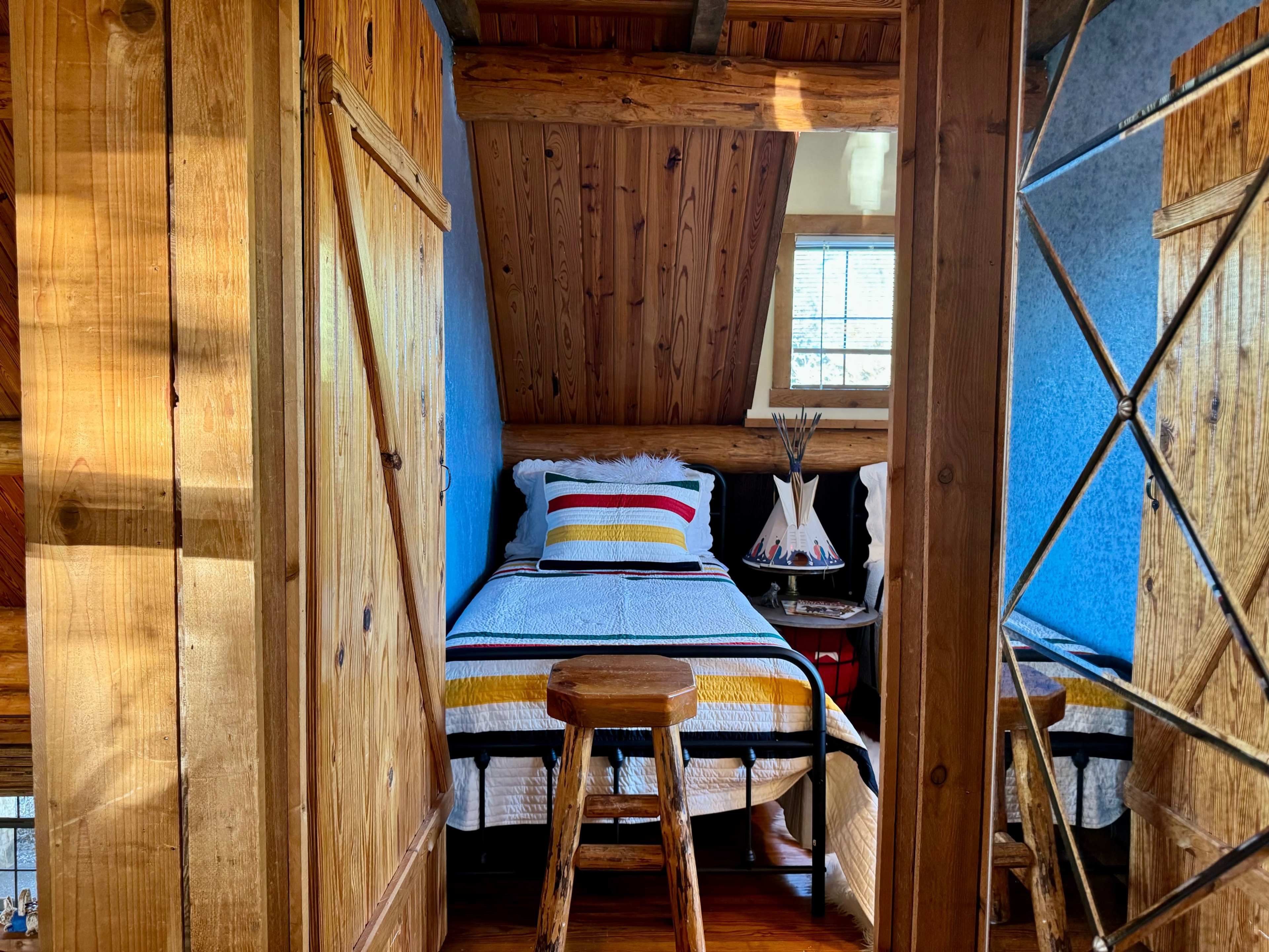 A cozy bedroom features a single bed with a striped blanket, wooden decor, and a small stool, set in a cabin-like environment.