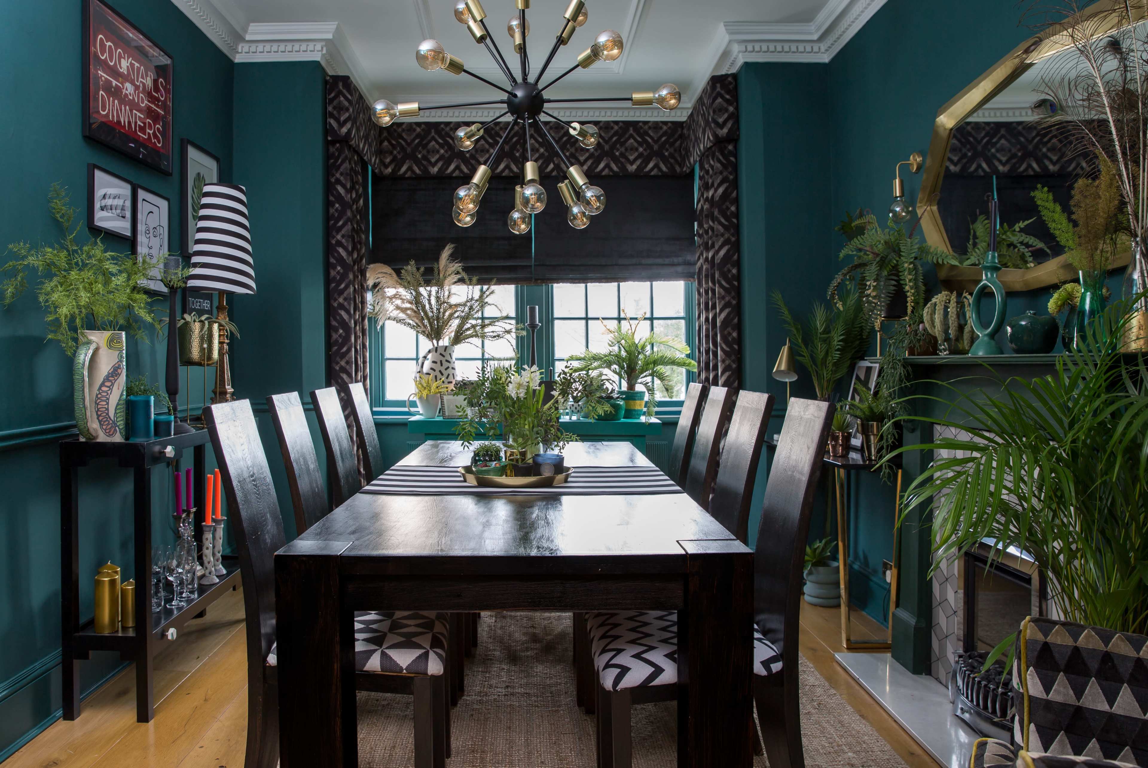 A large dining table with several chairs is positioned in a room adorned with green walls, plants, and modern lighting fixtures.