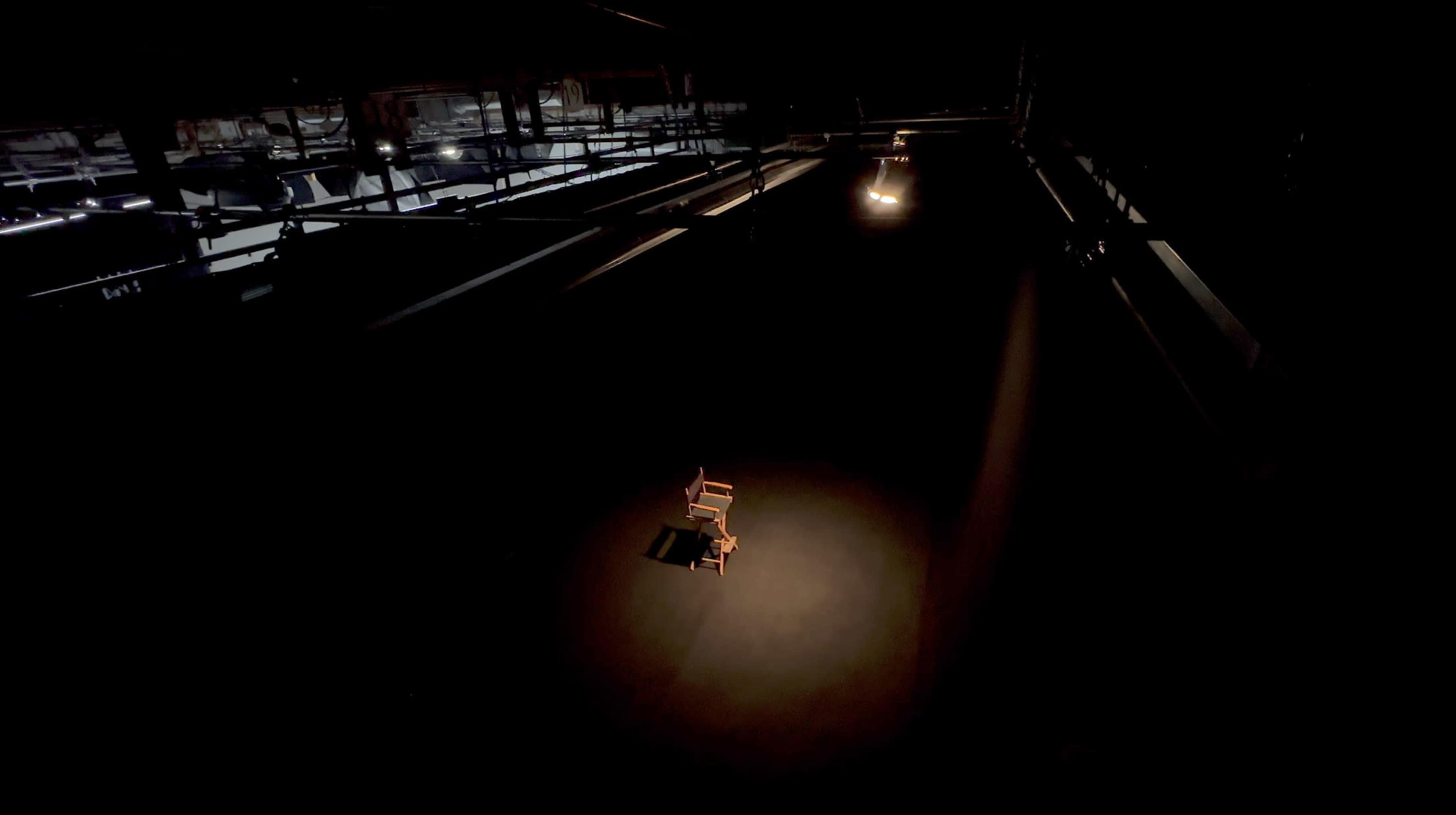 A single wooden chair is illuminated by a spotlight in an otherwise dark warehouse.