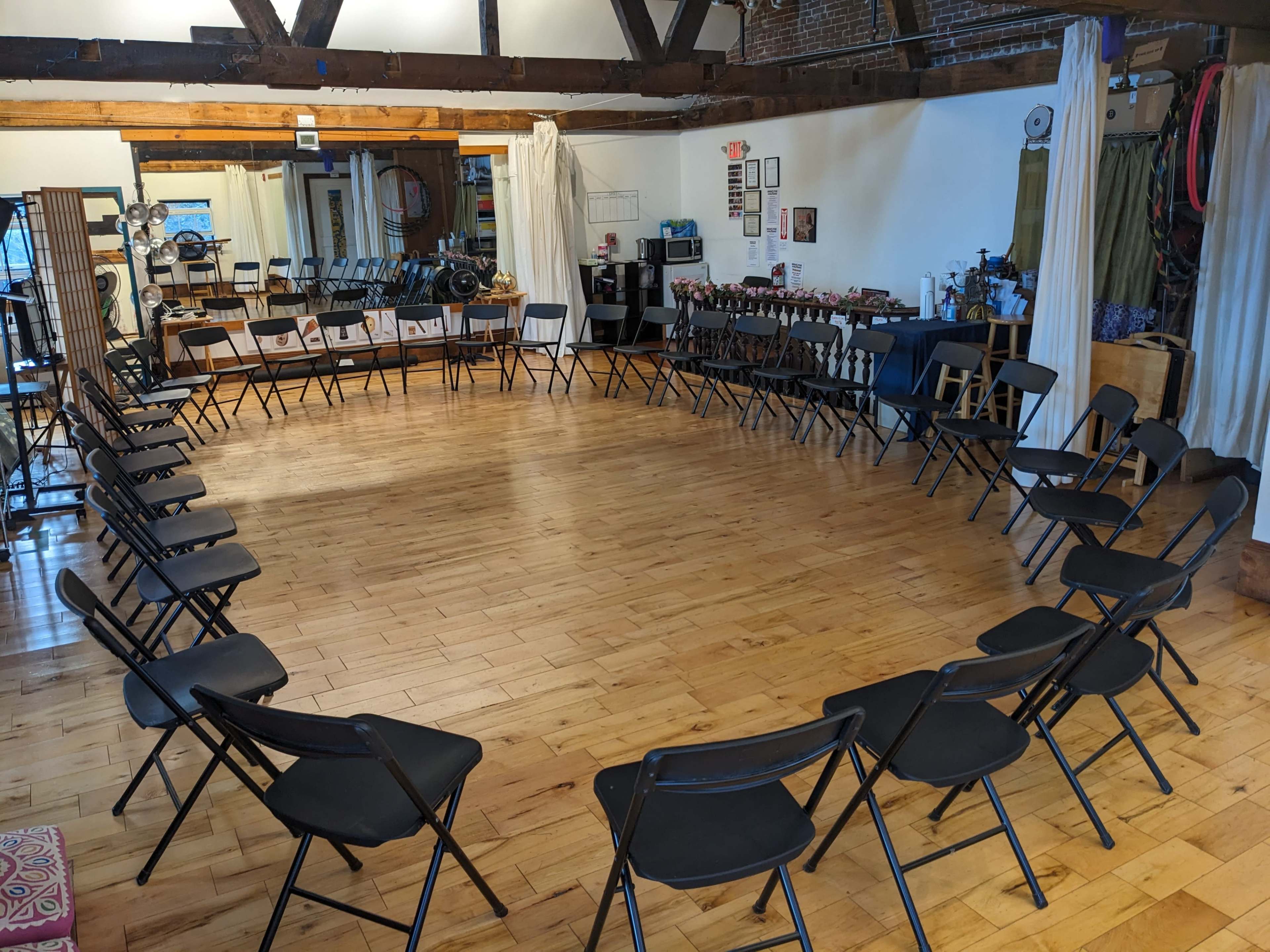 A spacious room with a circular arrangement of black folding chairs on a wooden floor, surrounded by various items and decorations along the walls.